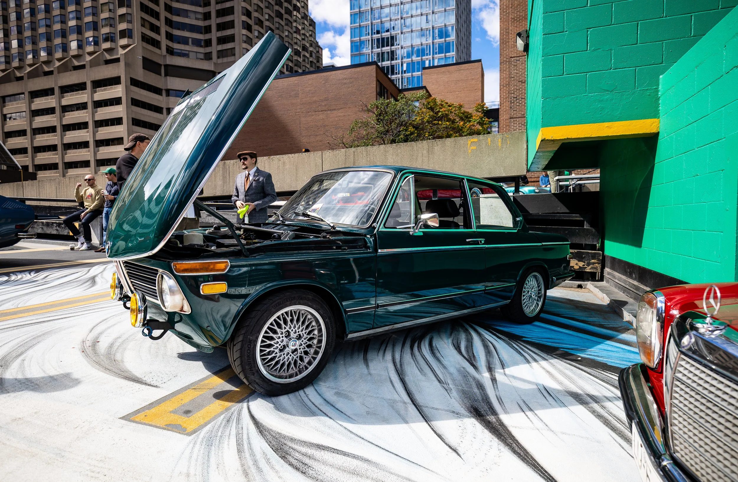 A vintage green car with its hood open parked on a city street during the day, with people and tall buildings in the background.