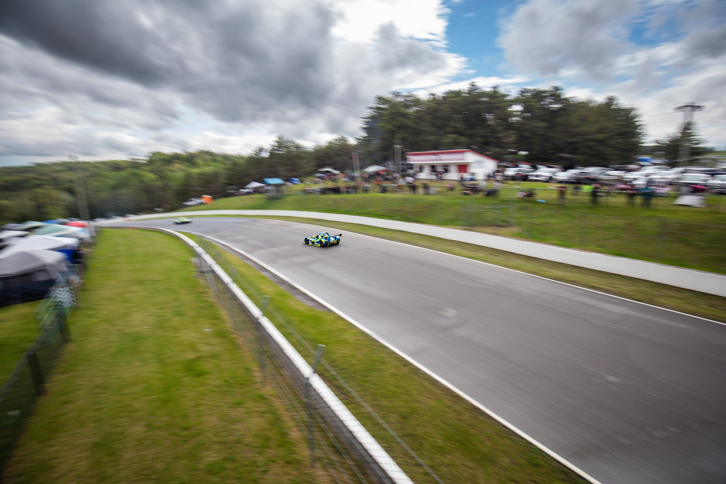 Race cars speeding on a curved race track with a crowd watching from the sidelines under cloudy skies.