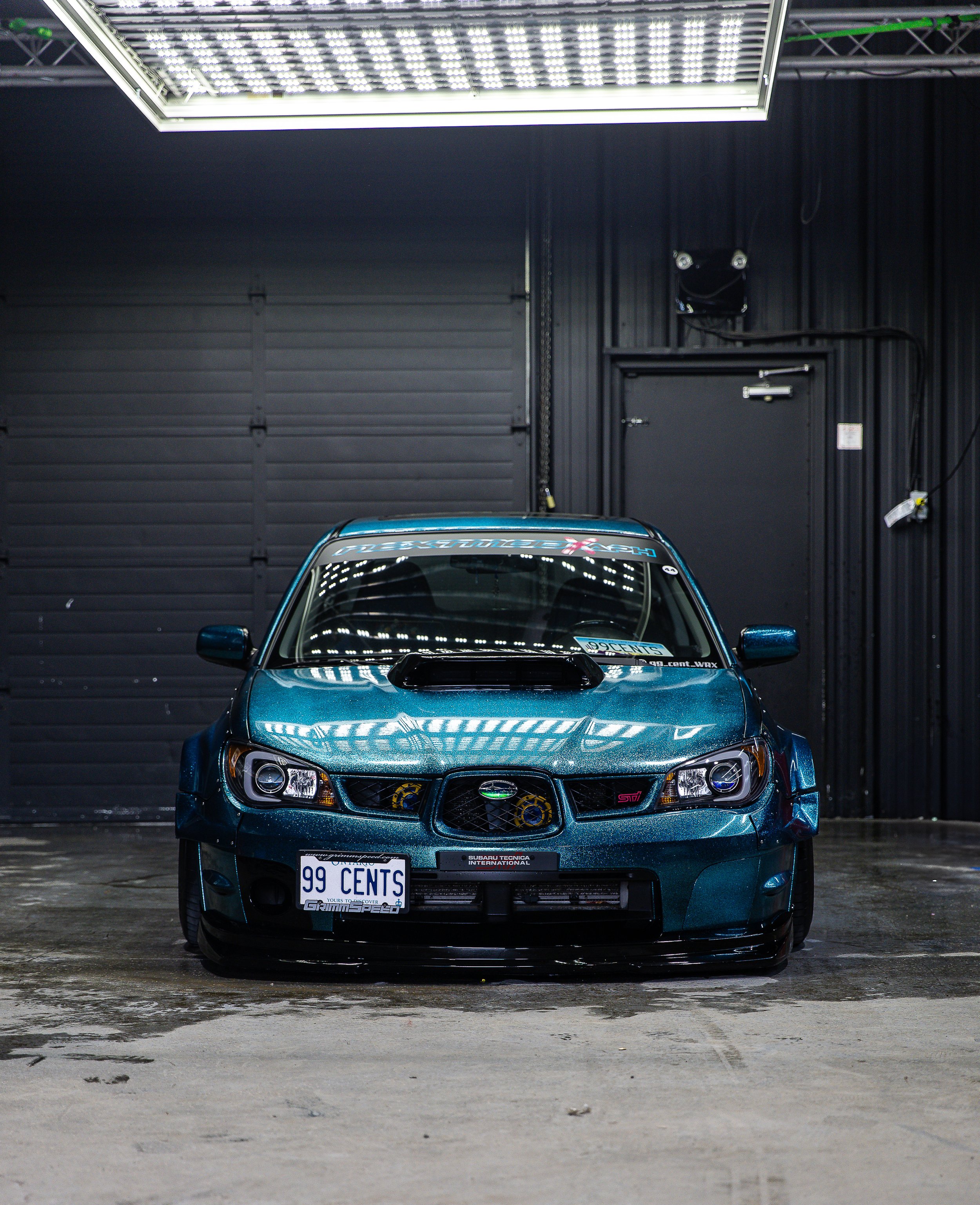 A blue Subaru WRX car parked inside a garage with black walls, partially visible from the front, with a license plate reading '99 CENTS' and various stickers on the windshield.
