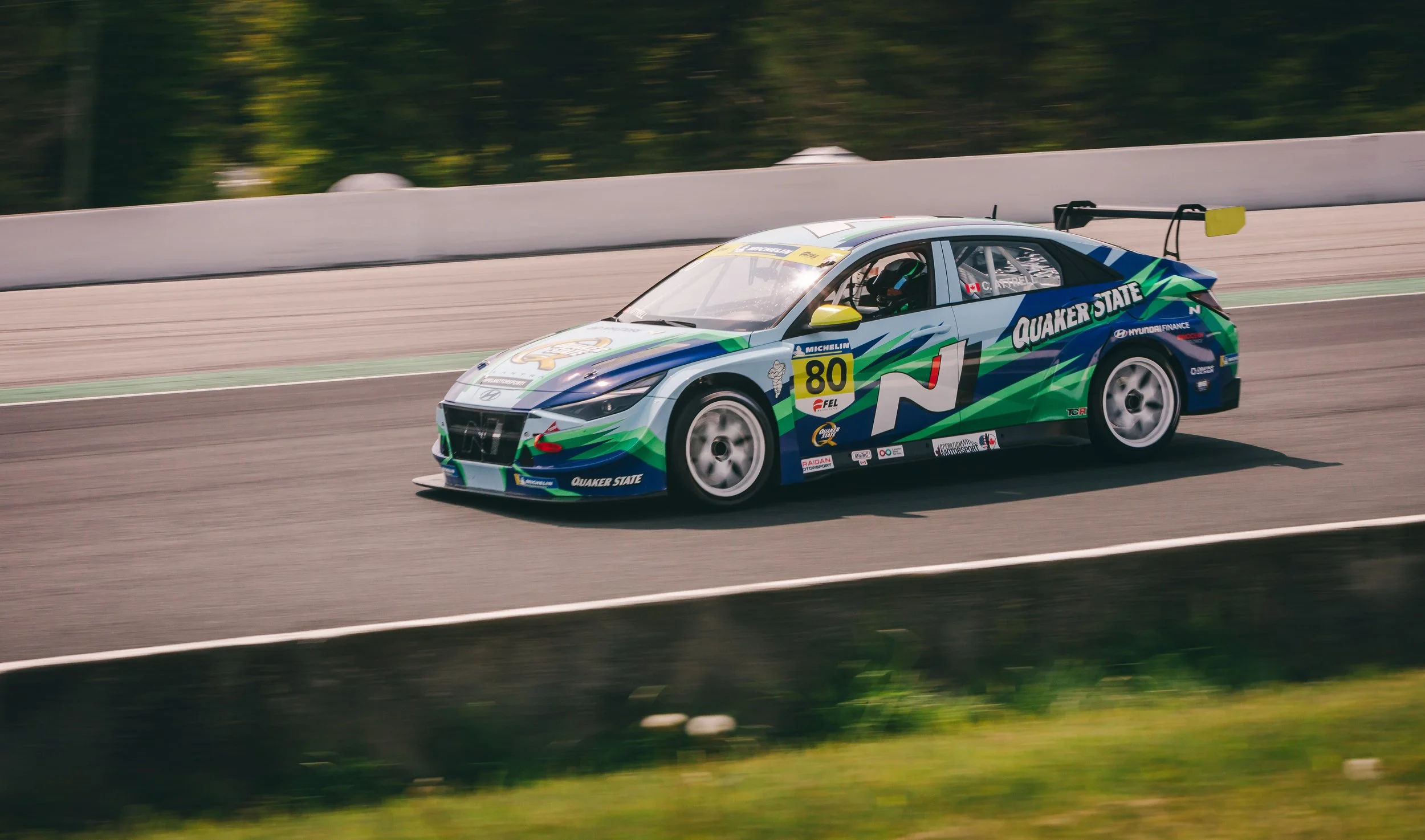 A race car speeding on a track, decorated with green, blue, and white graphics and the words 'Quaker State' on the side.