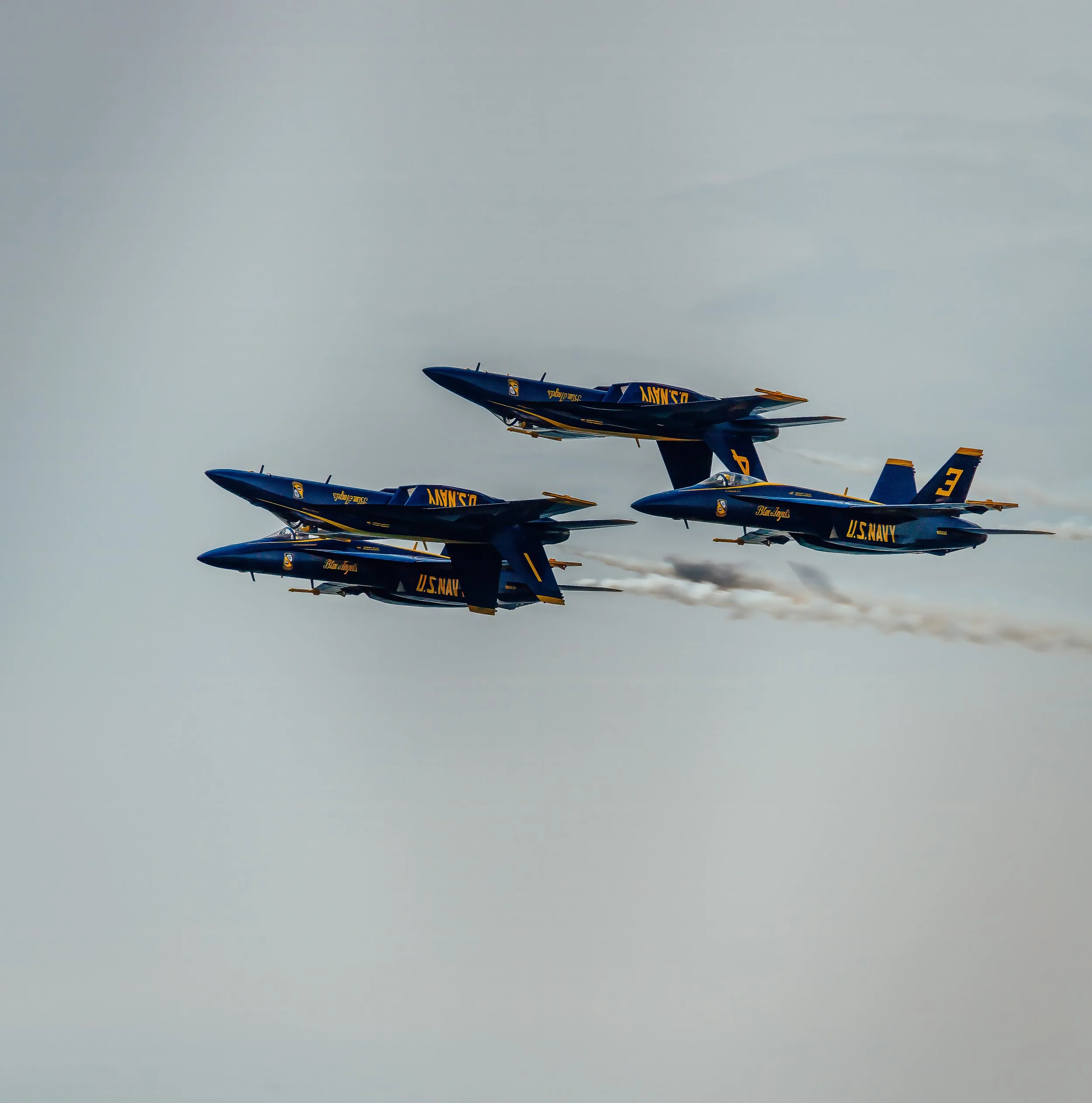 A formation of five Blue Angels fighter jets flying in close formation against a cloudy sky, with blue and yellow military markings and the US Navy insignia.