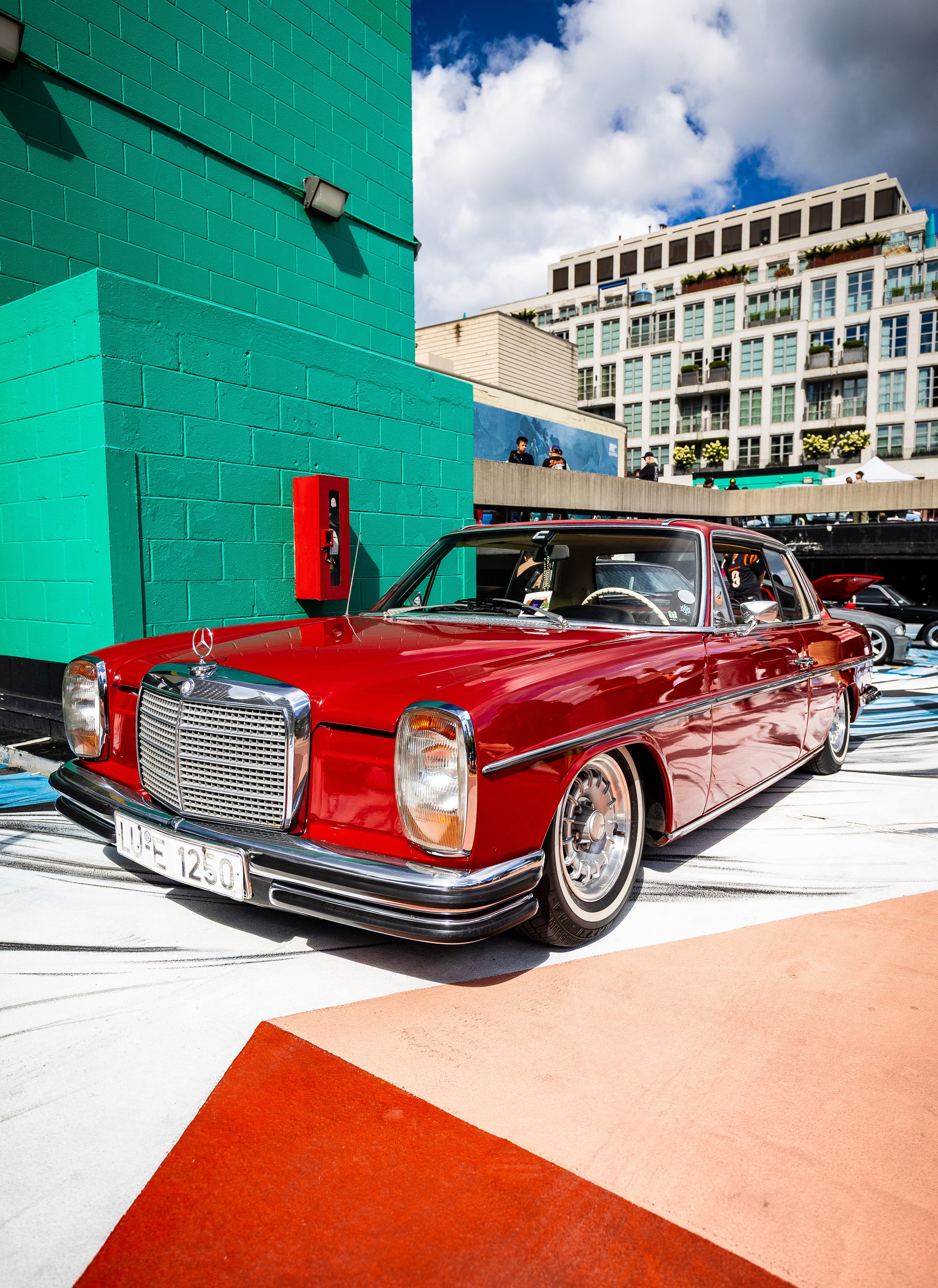 A vintage red Mercedes-Benz car parked in a colorful urban setting with a teal green building on the left and a modern white residential building in the background, under a partly cloudy sky.
