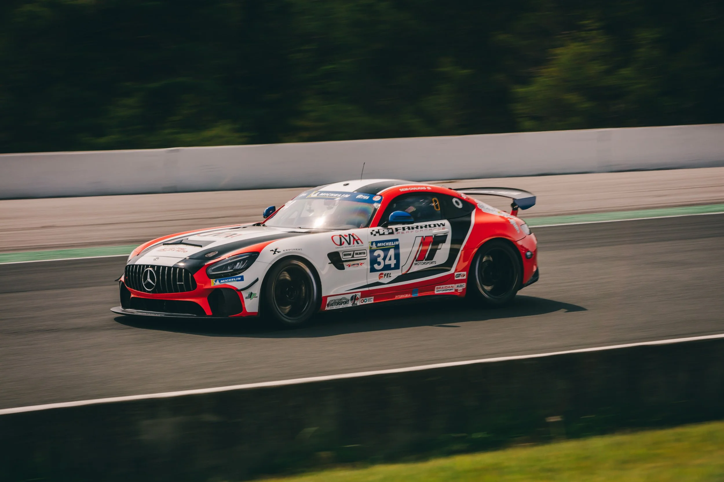 A racing car on a race track, featuring a red, white, and black color scheme with various sponsor logos including Mercedes-Benz, Michelin, and others. The car is moving at high speed with motion blur in the background.