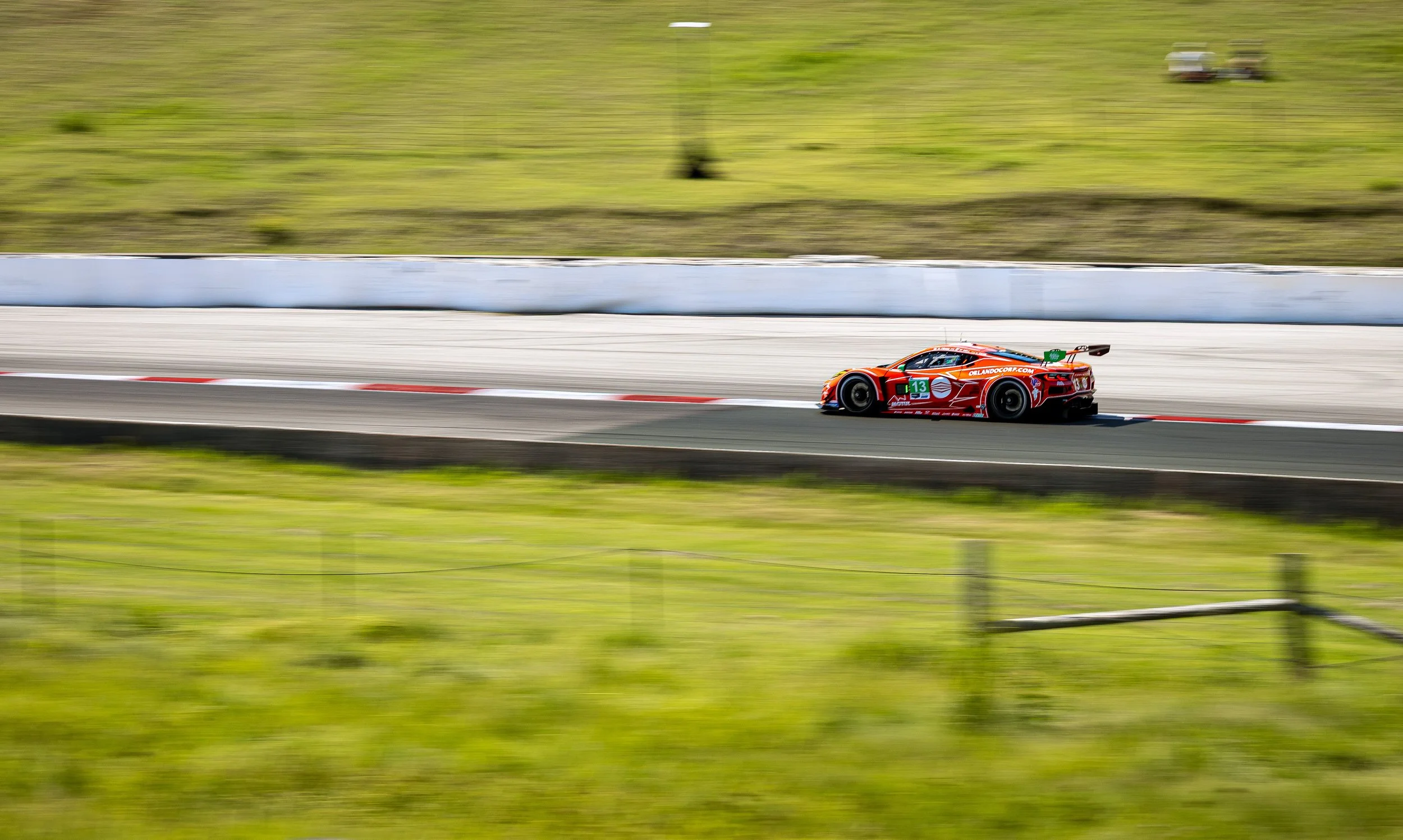 A red racing car with the number 13 on its side driving at high speed on a race track, with a blurred background of green grass and trees.