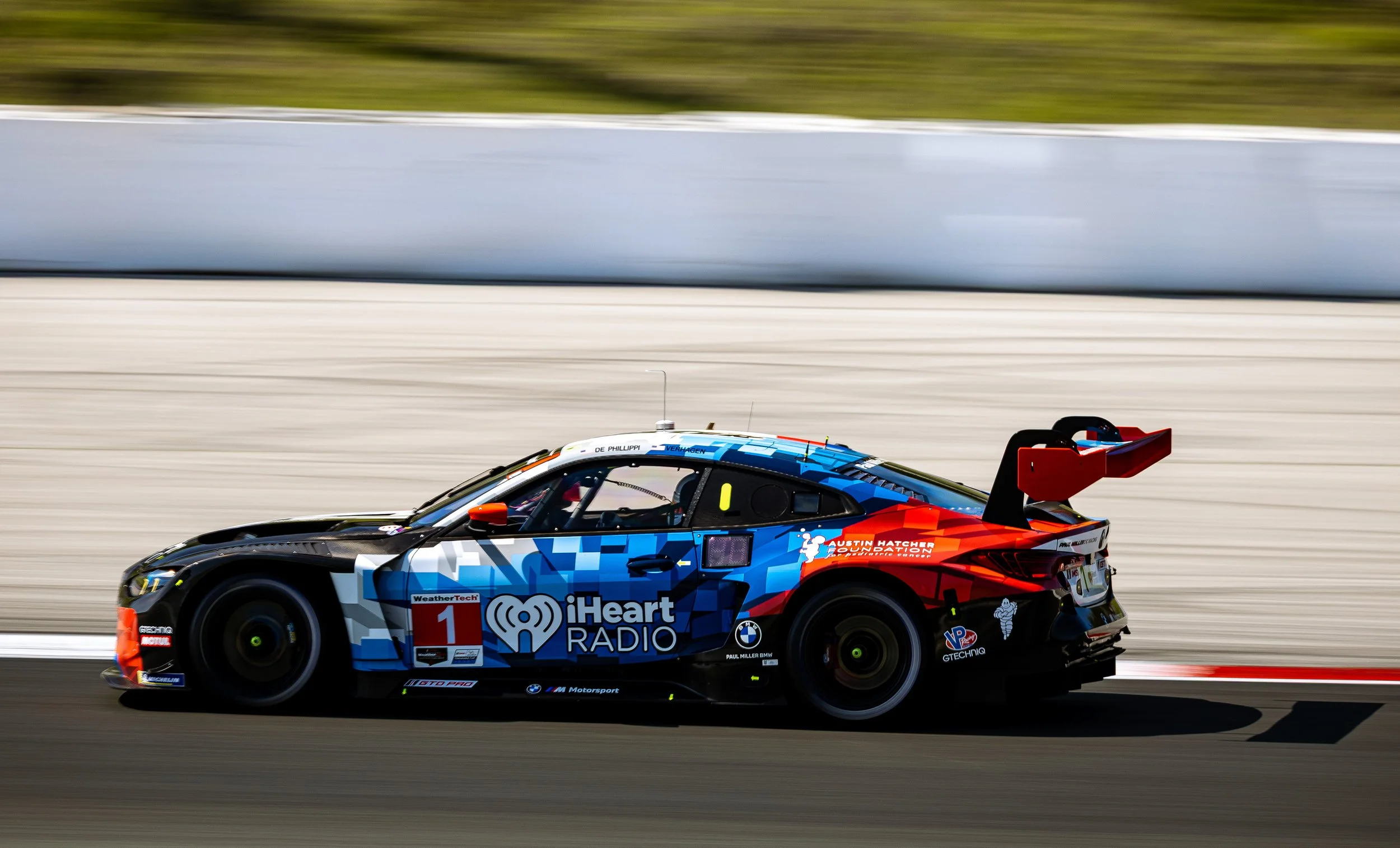 A race car with a blue and black design, sponsorship logos including iHeart Radio and Austin Hatcher Foundation, speeding on a race track with motion blur in the background.