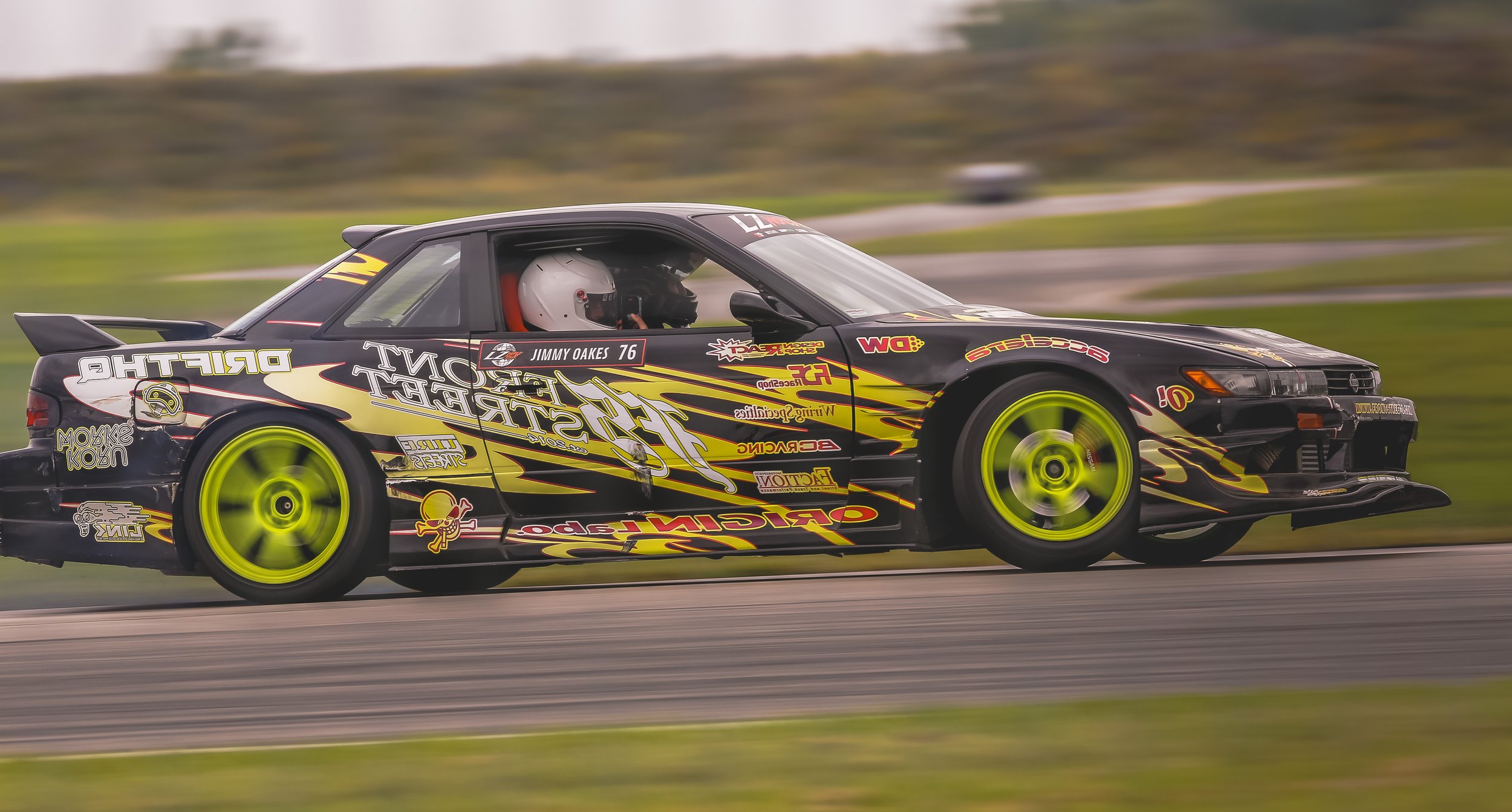 Race car with bright yellow wheels and colorful sponsor decals speeding on a track, driver wearing white helmet inside.