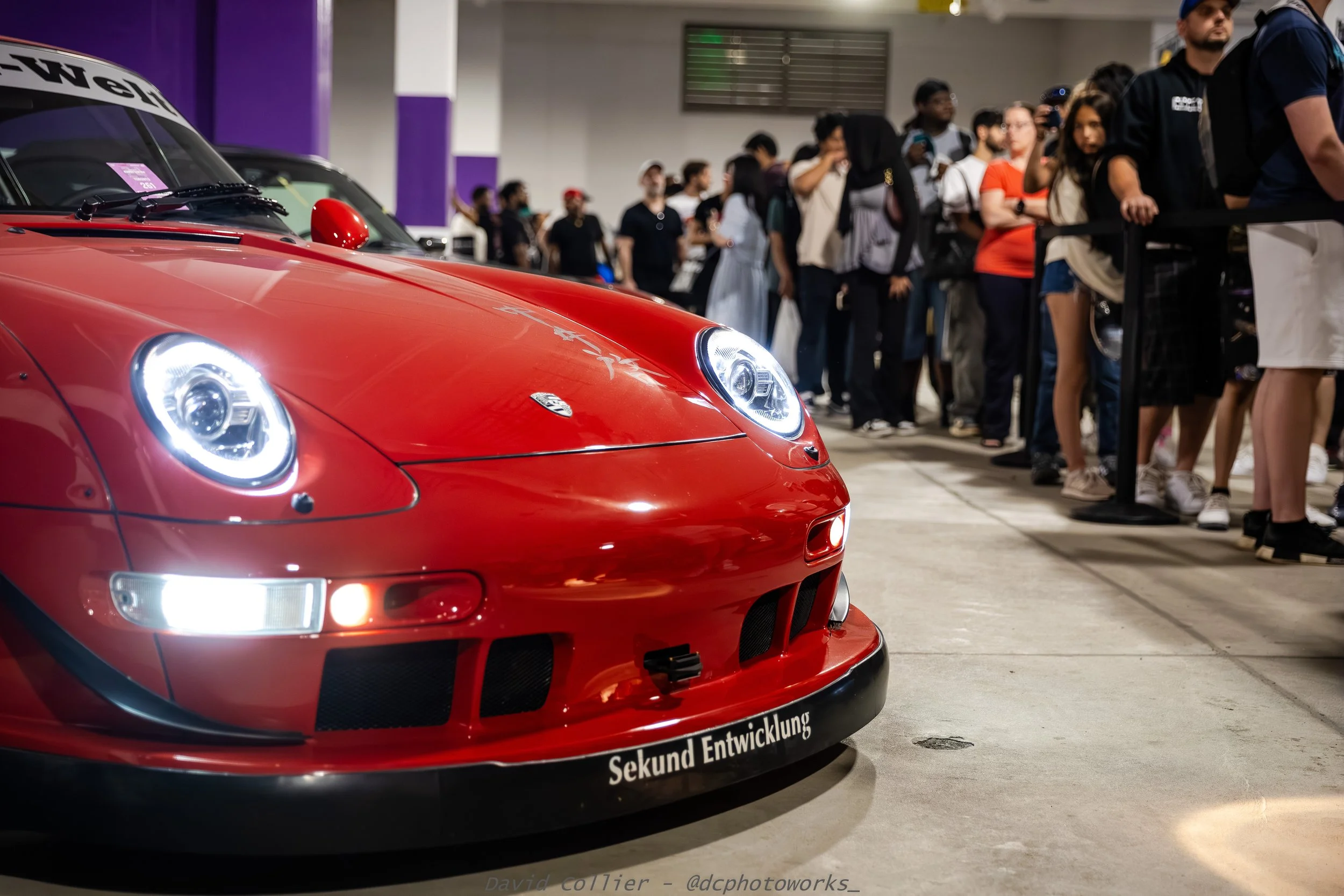 Close-up of a red sports car with headlights on, parked at an indoor car show with a line of people in the background.