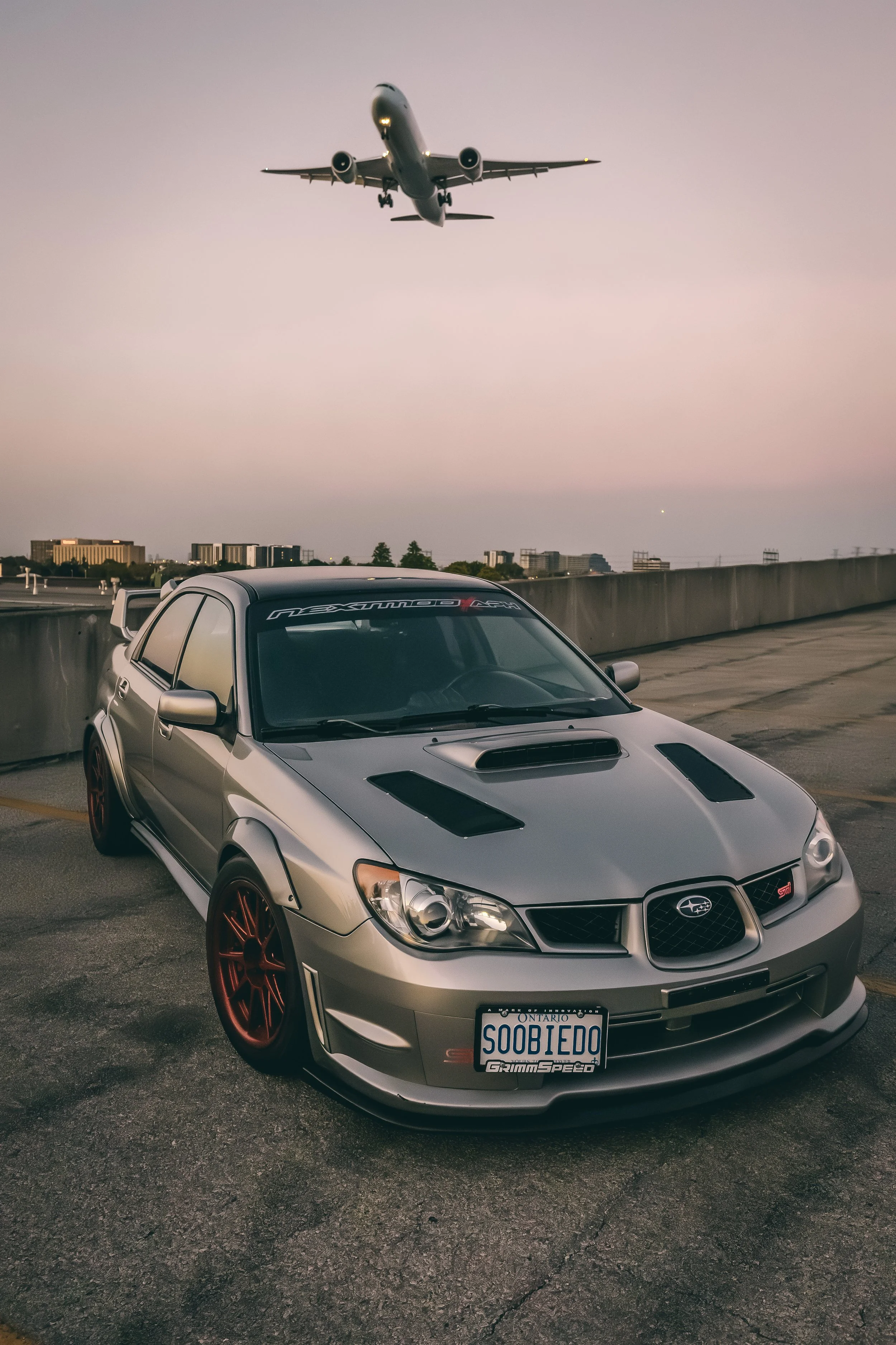 A silver Subaru WRX car with red wheels parked on a rooftop parking lot with a gray sky in the background. An airplane is flying low overhead. The car has a custom Ontario license plate reading "SODBENTO" and a GrimSpeed sticker on the front windshie