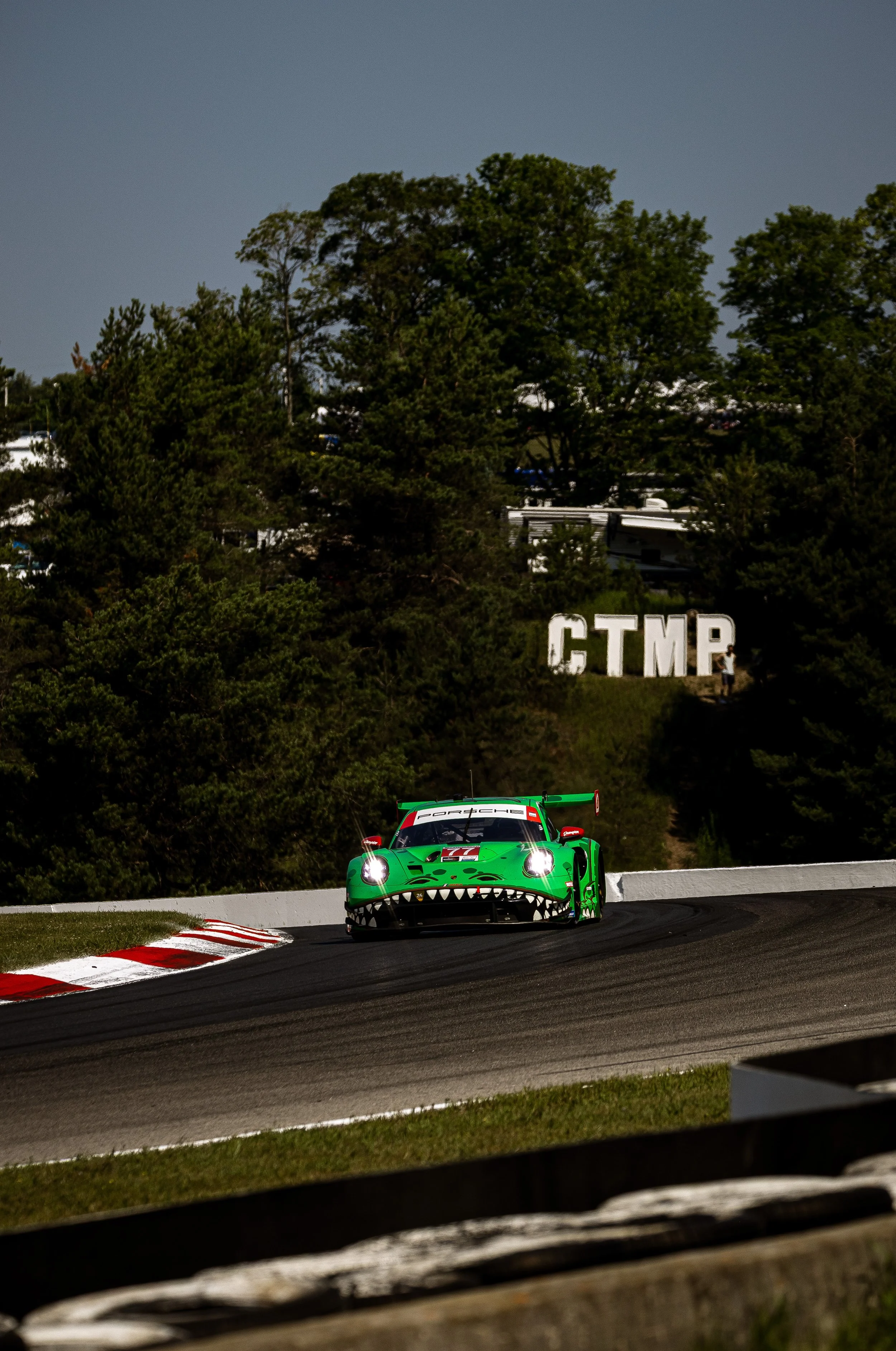 A green race car with a dinosaur mouth design on the front, racing on a track with a backdrop of trees and a sign that reads 'CTMP' in large white letters.