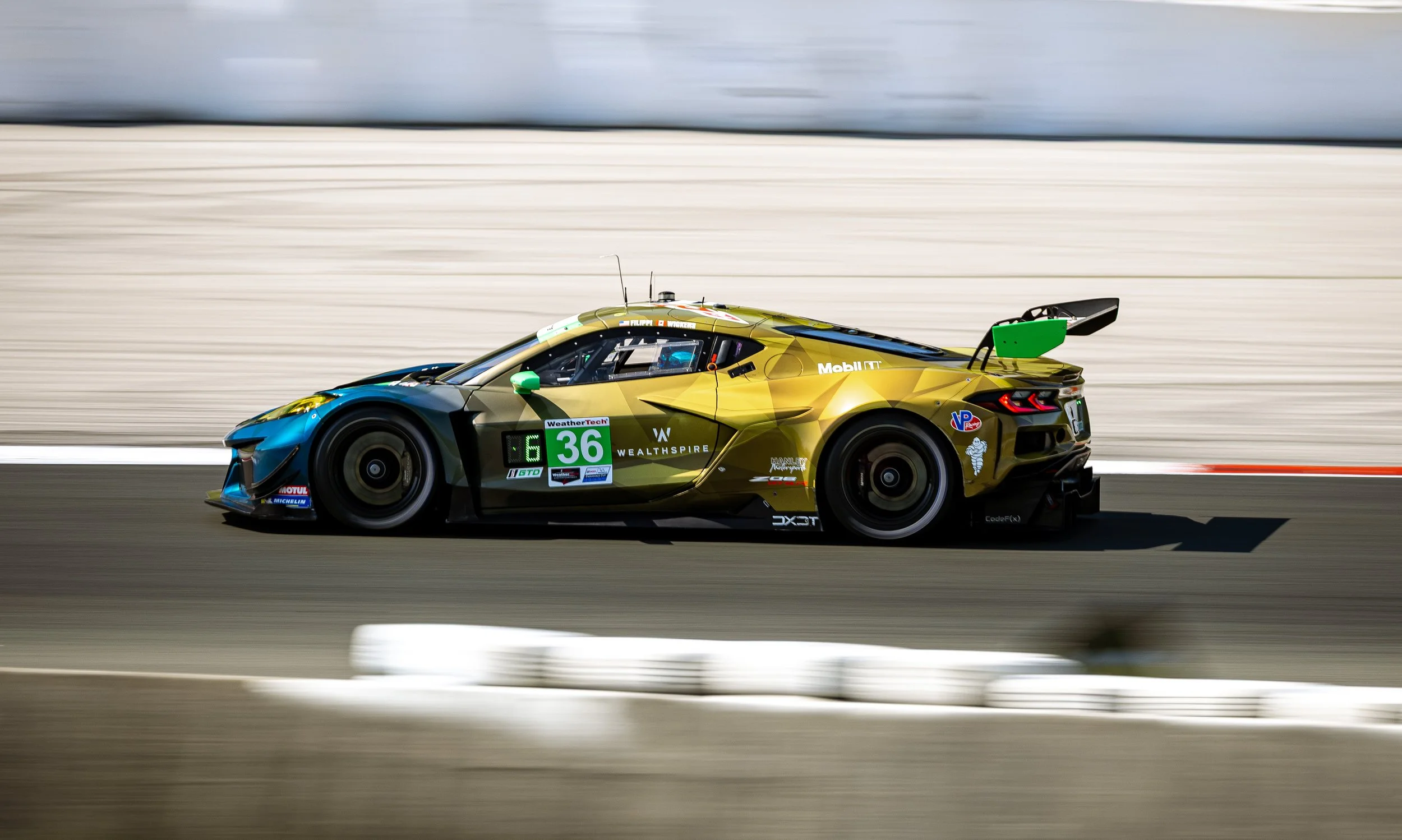 A race car driving on a track, with motion blur background, featuring a yellow, blue, and black color scheme, and various sponsor logos, including Michelin, WeatherTech, and Wealthspire.