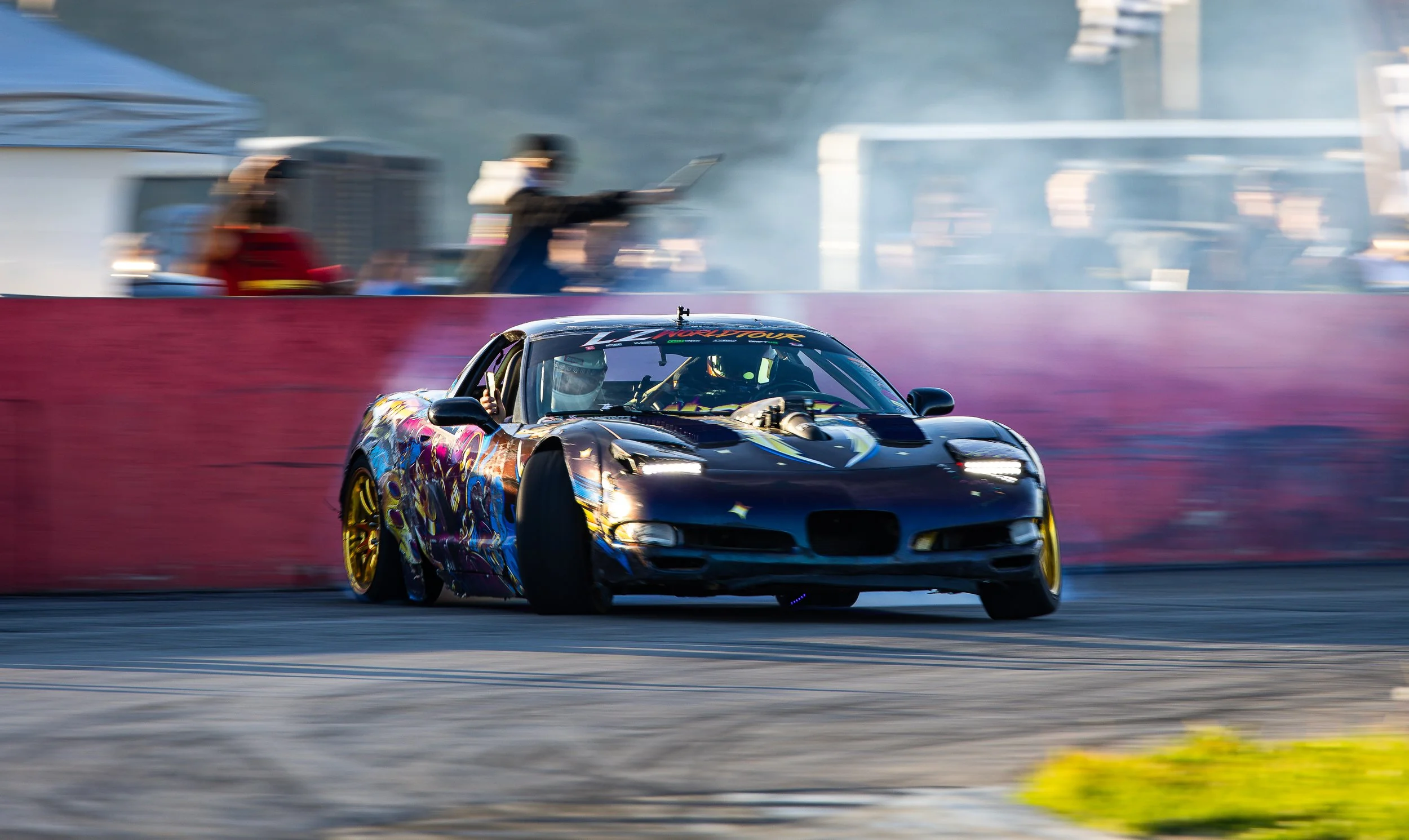 A sports car with a colorful, graphic design on its body racing on a track, with blurred spectators and officials in the background.