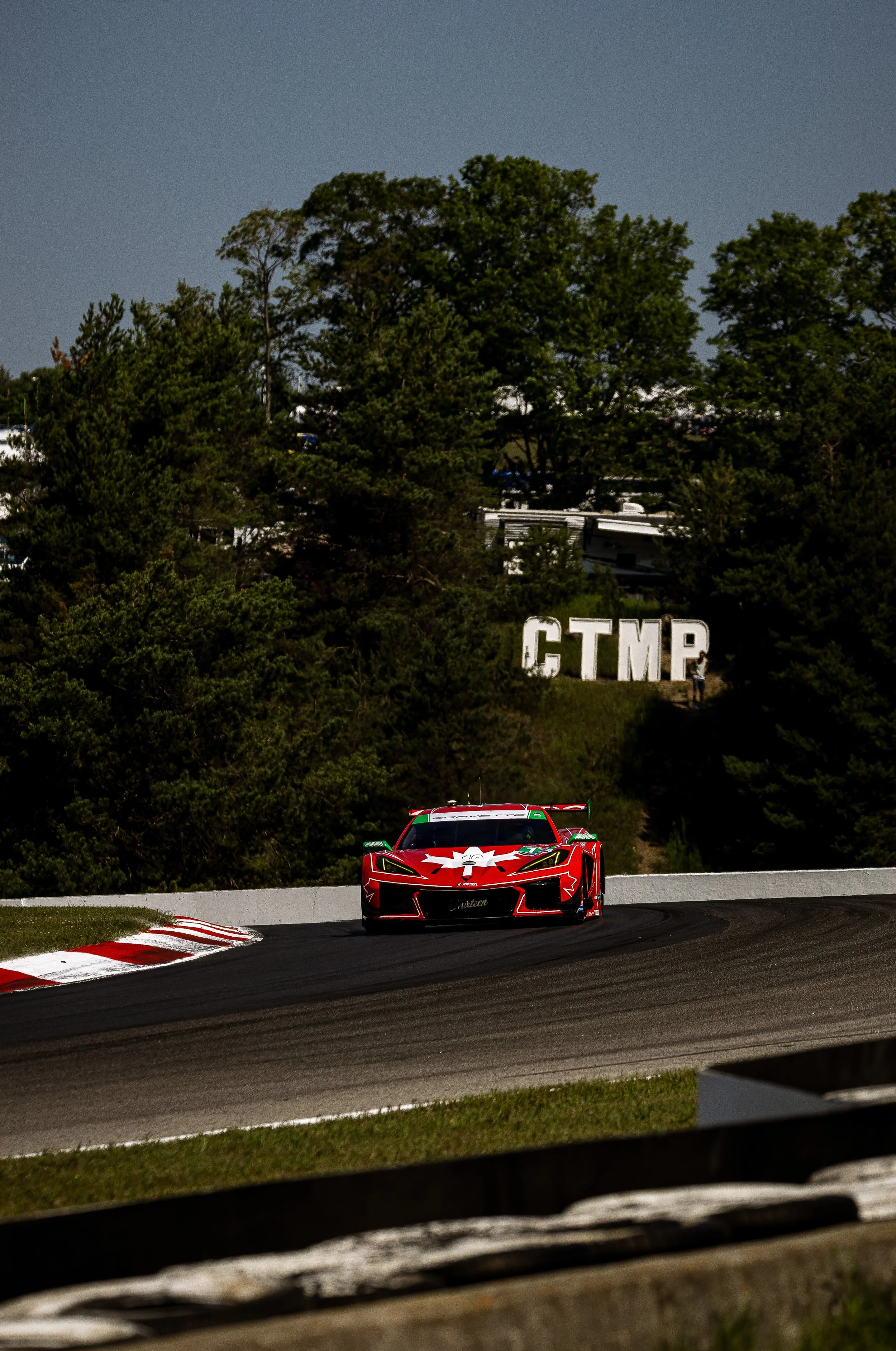 A red race car is driving on a race track surrounded by trees, with a large sign that reads 'C.T.M.P.' in the background, and a person standing nearby.