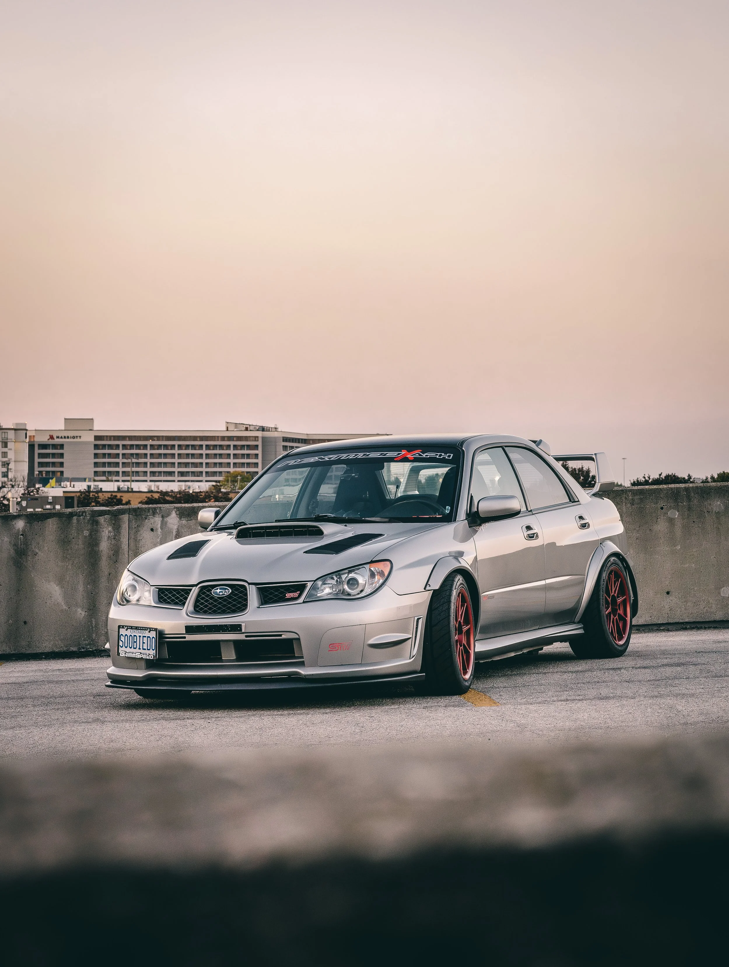 A silver Subaru Impreza WRX STI parked in an outdoor lot with a cityscape background under an overcast sky.