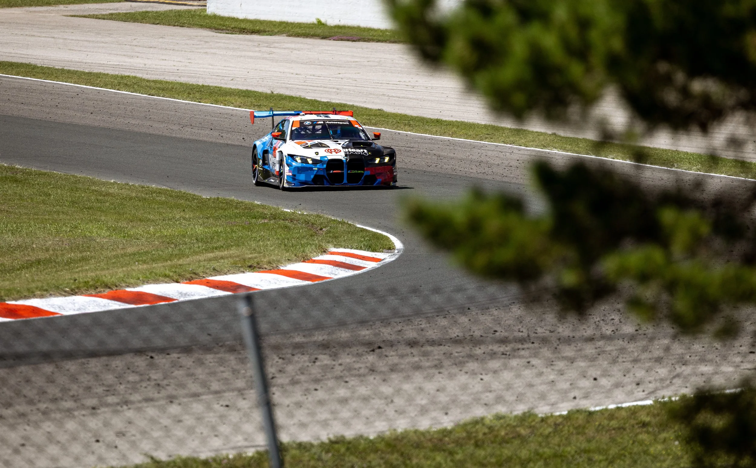 A race car on a racing track, seen through some greenery, with a white wall and grassy area in the background.