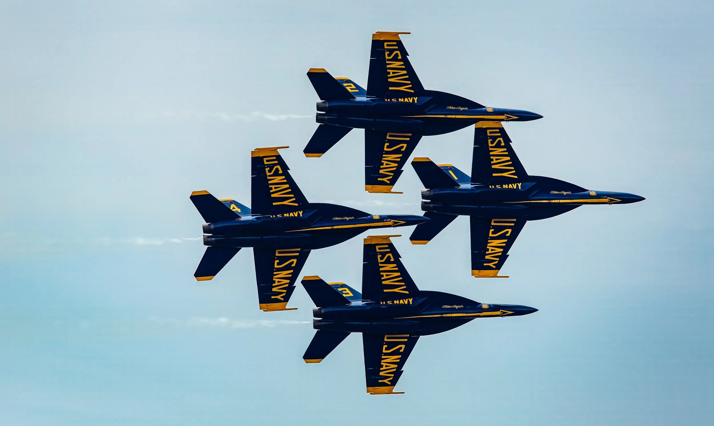 Blue Angels fighter jets flying in formation, with U.S. Navy markings, against a light blue sky.