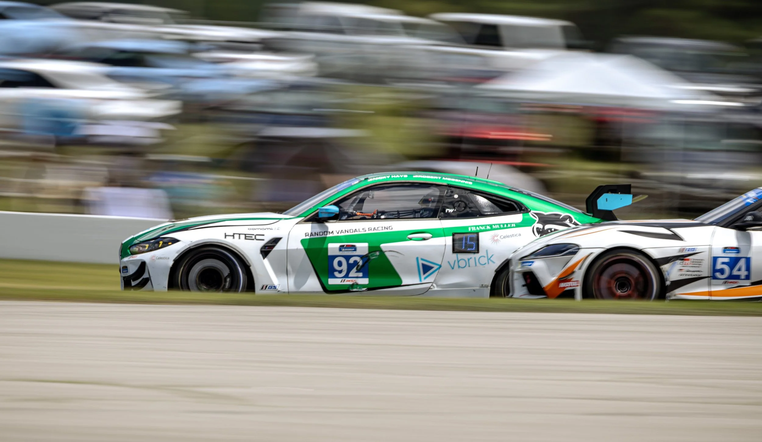 Blurry race car with the number 92 racing on a track, with a mostly white body, green accents, and sponsor logos, passing another car and a background of multiple parked cars.