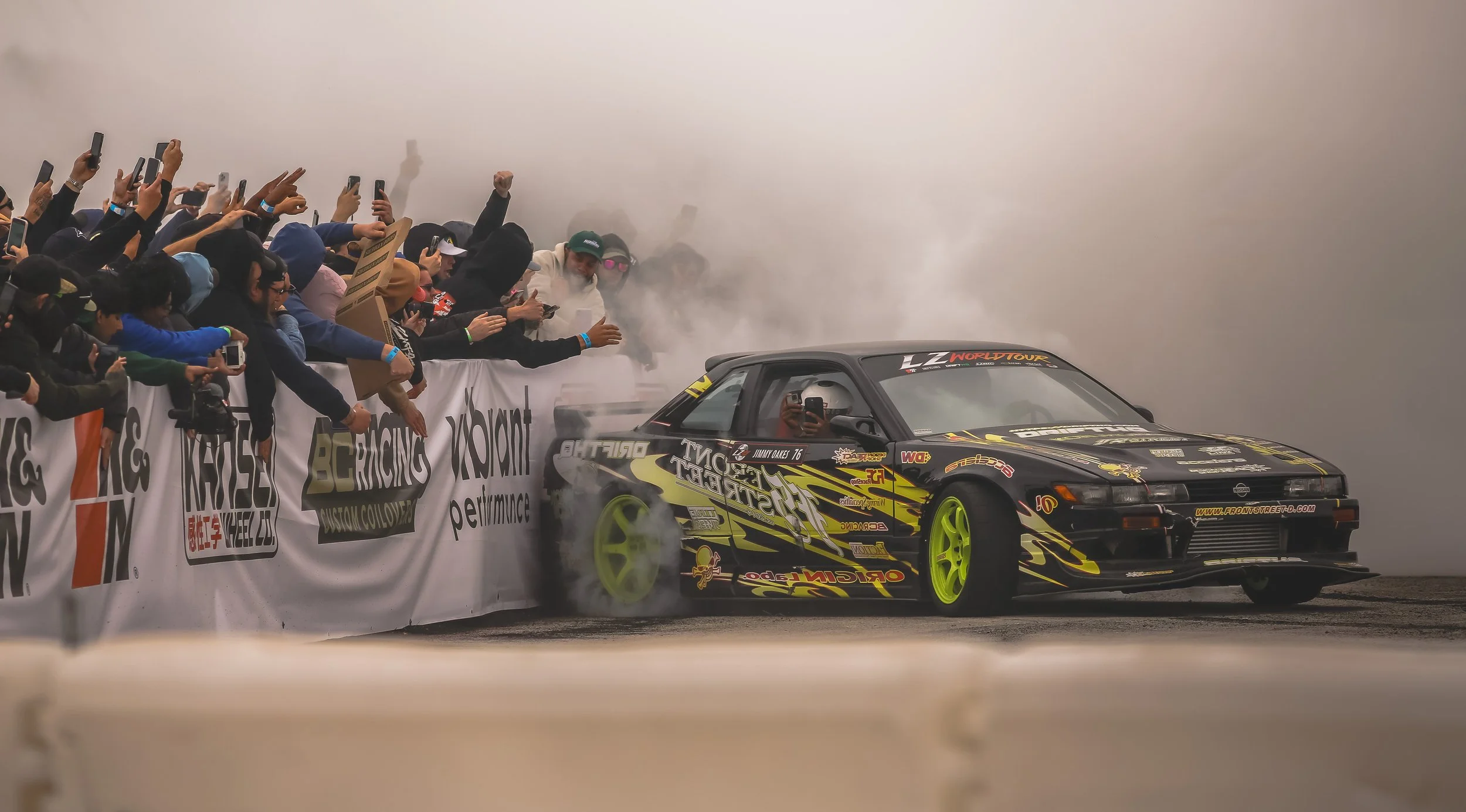 Race car producing burnout smoke as it performs at a motorsport event, with spectators taking photos and cheering from behind a barrier.