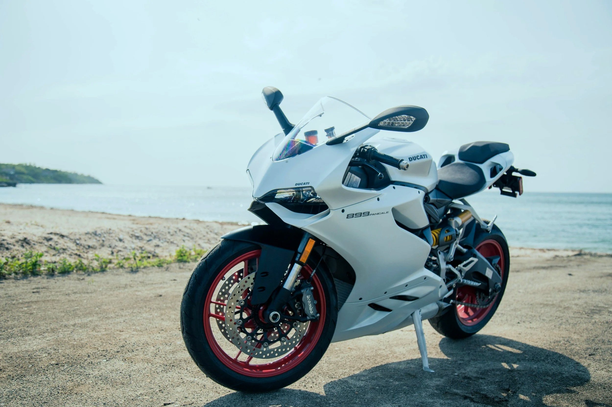 A white Ducati Panigale motorcycle with red wheels parked on sandy beach near water under a cloudy sky.