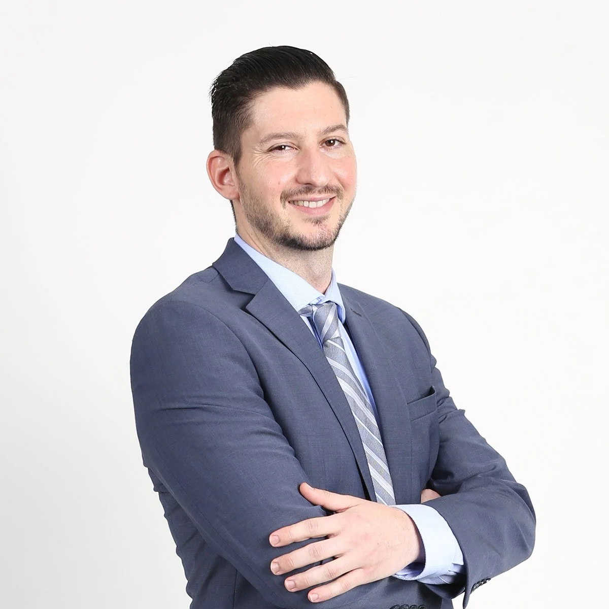 A young man in a dark blue suit with a light blue dress shirt and striped tie, smiling with his arms crossed against a plain white background.