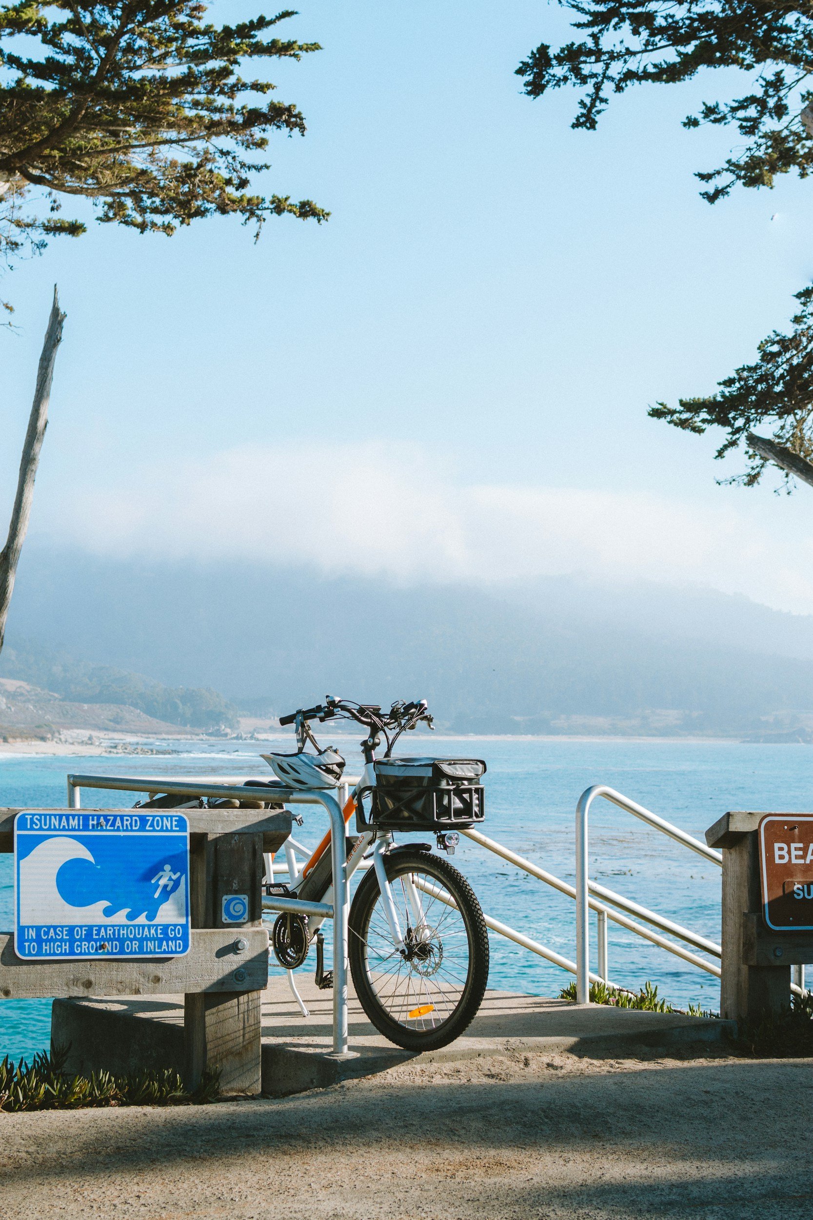 Beachside with a bicycle parked near a 'Tsunami Hazard Zone' sign, overlooking the ocean and distant shoreline with trees in the distance.