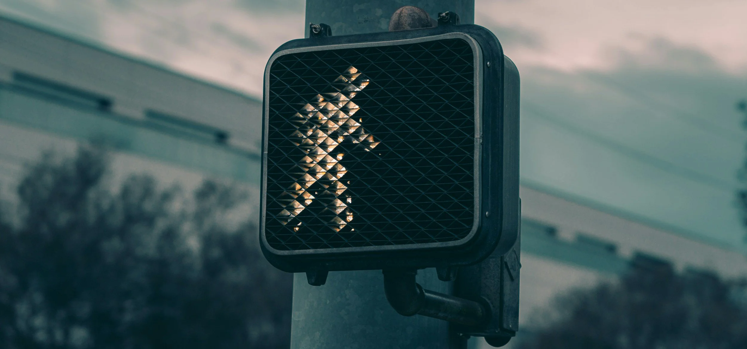A pedestrian crossing sign with a person walking, mounted on a pole outdoors, with blurred trees and building in the background.