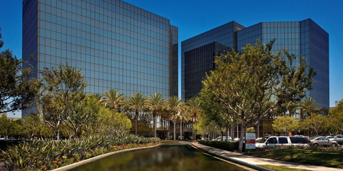 Two modern glass office buildings with reflections, surrounded by trees, palm trees, and a small water feature in the foreground on a clear sunny day.