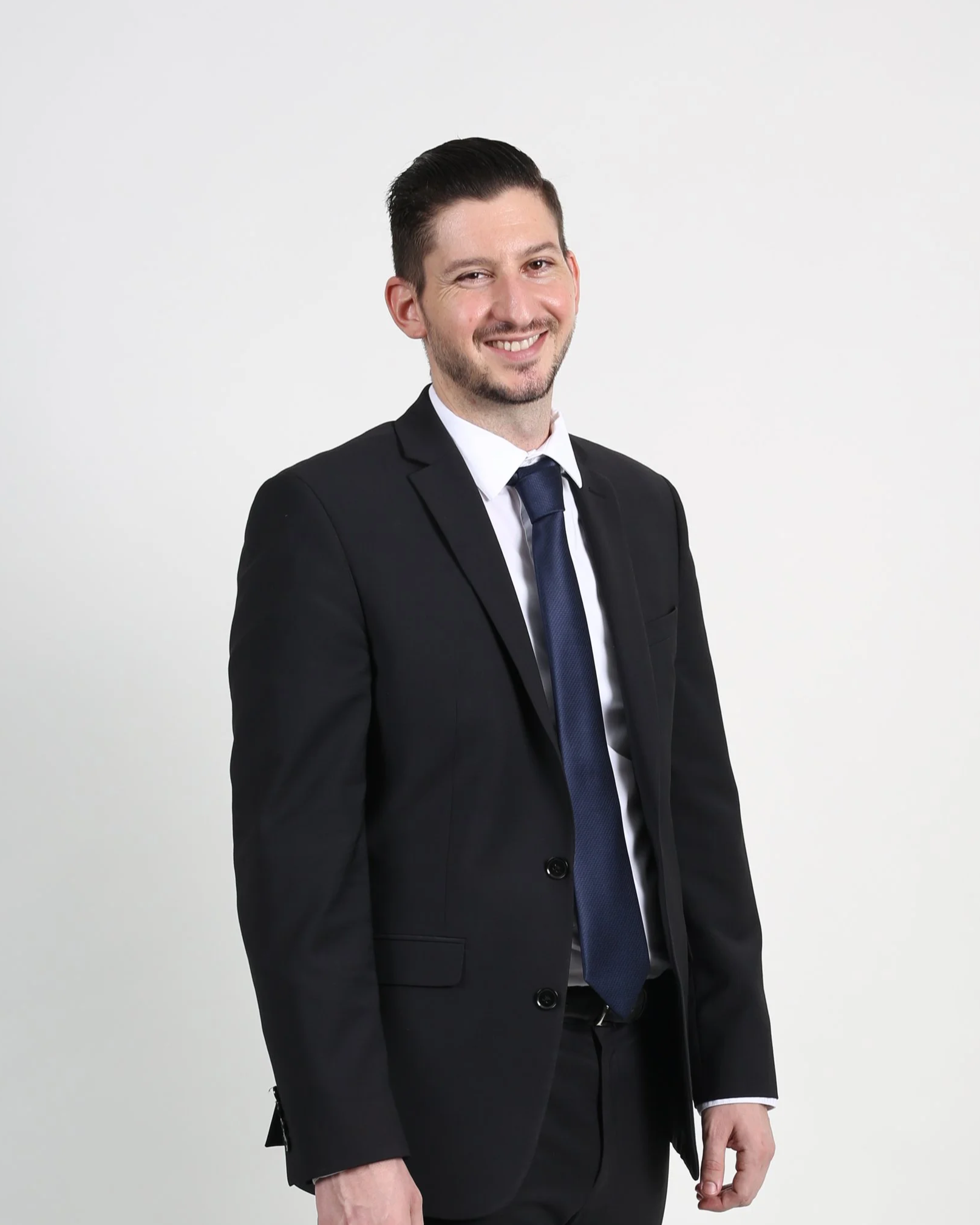 A man in a black suit, white shirt, and dark blue tie, smiling and winking, standing against a plain white background.