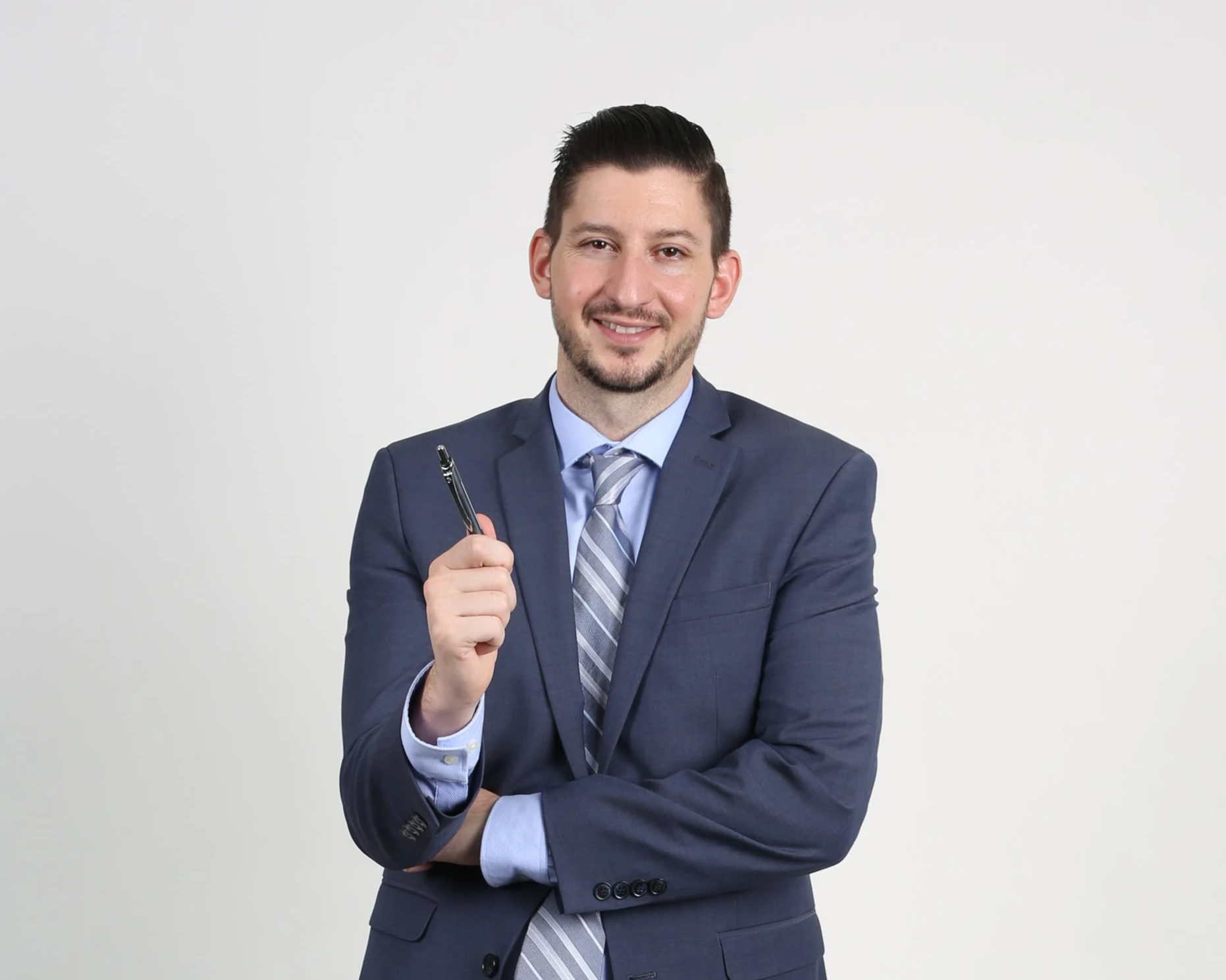 A young man in a blue suit with a striped tie, smiling and holding a pen, standing against a plain white background.