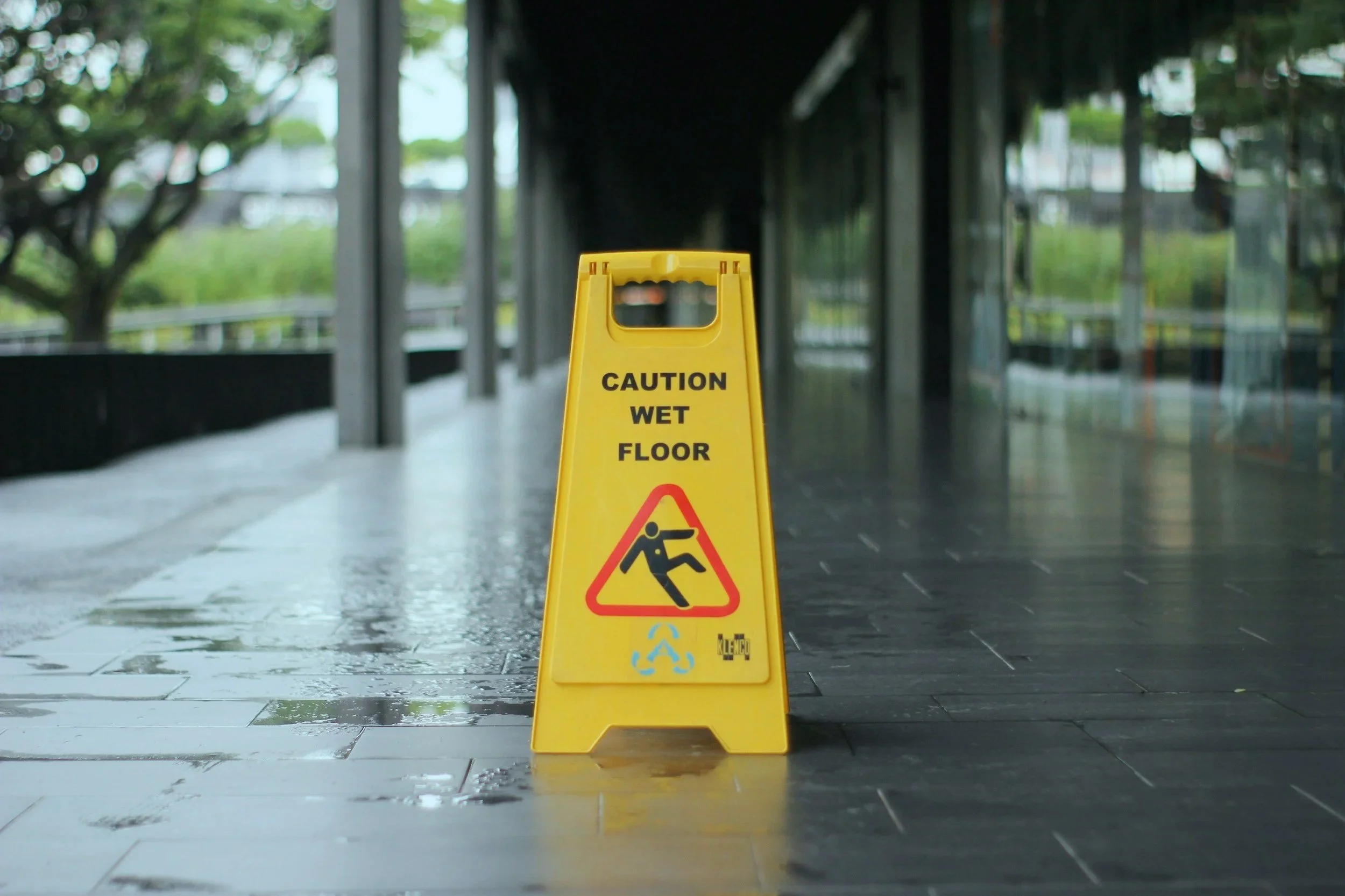 A yellow caution wet floor sign on a shiny, wet tiled floor in an indoor area, with glass walls and trees visible outside.