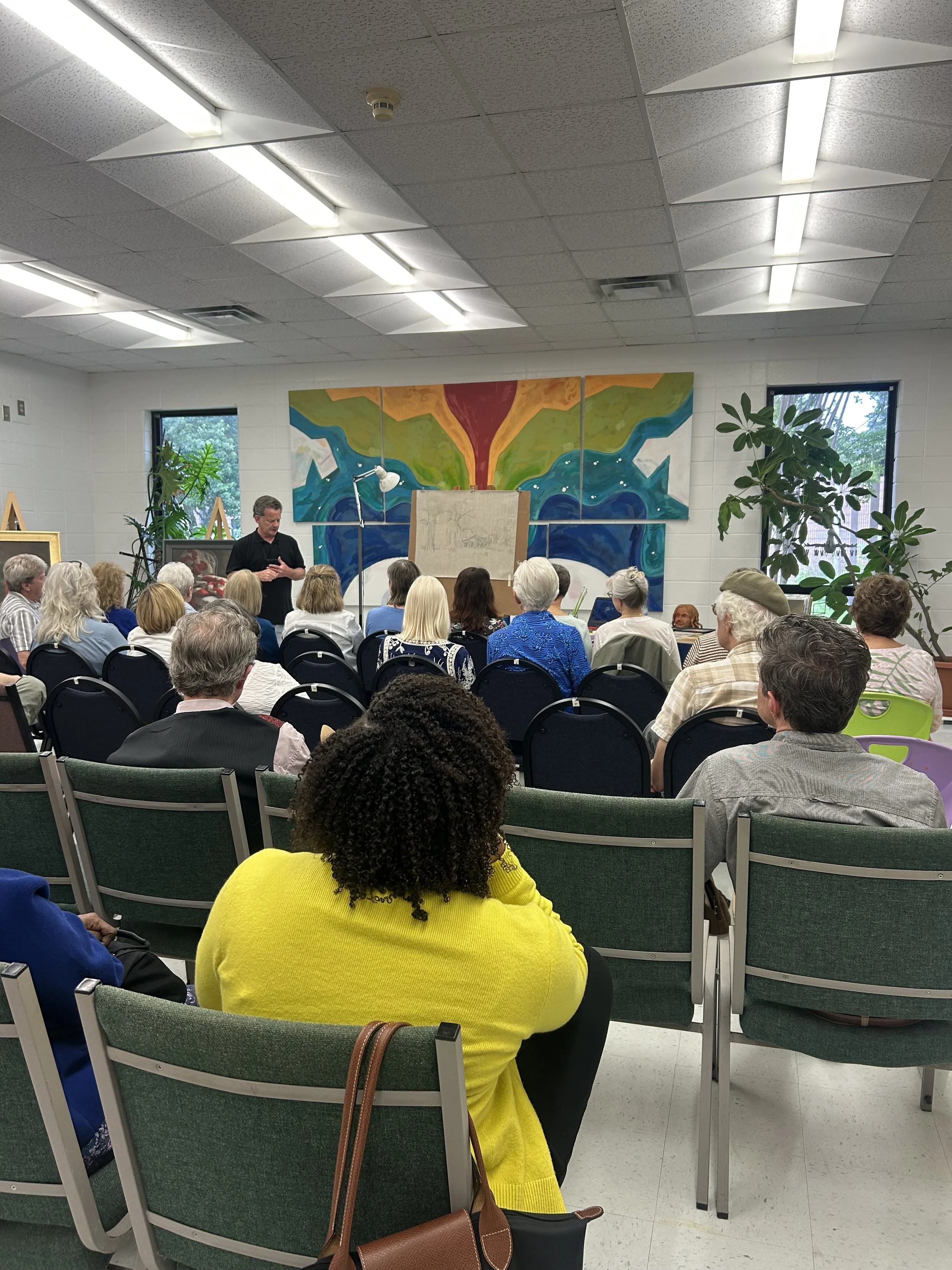 A group of people sitting in a room attending a presentation. A man is standing by a large painting on the wall.