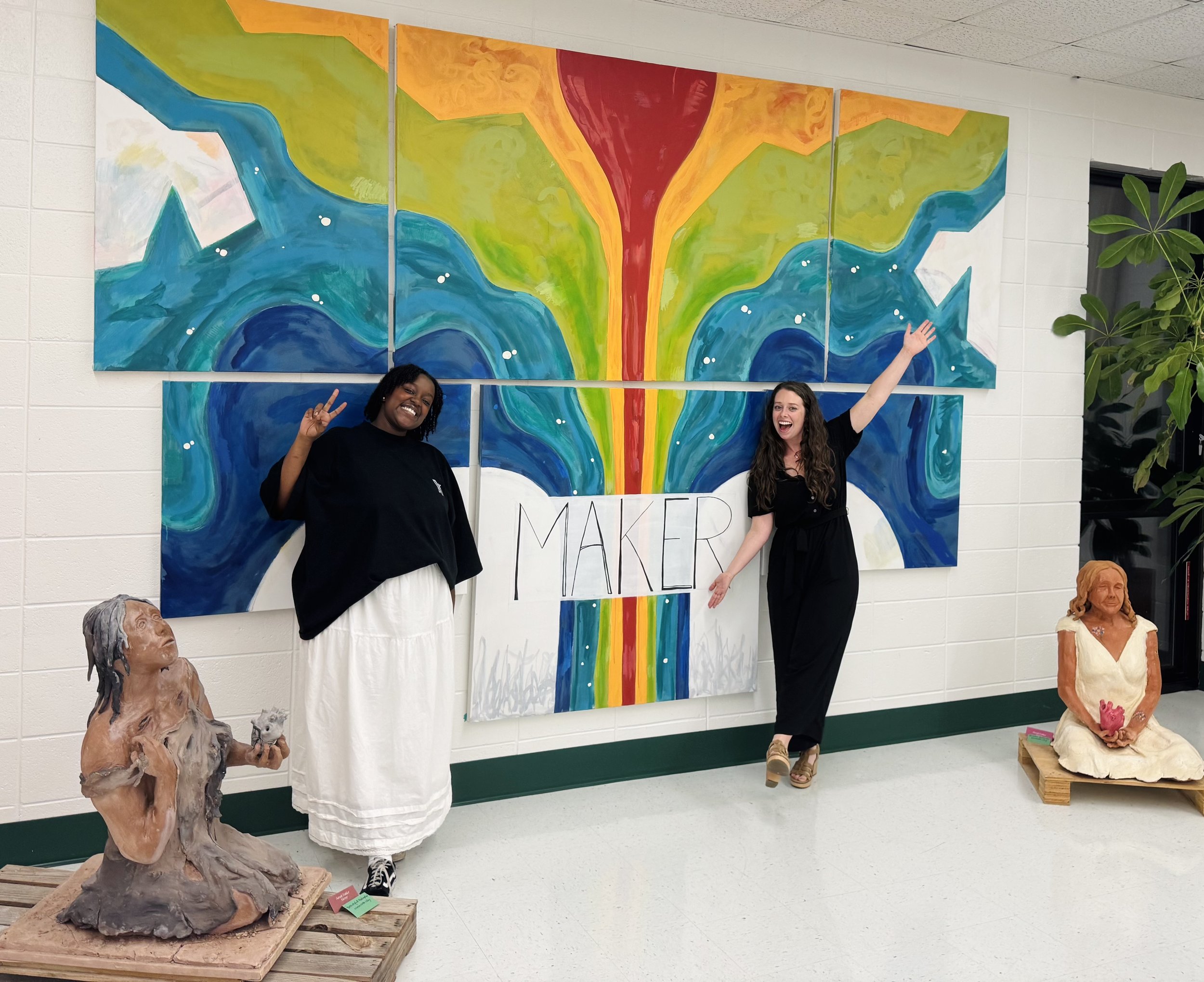 Two women are standing in front of a colorful abstract mural with flowing green, yellow, orange, red, and blue shapes, and a large red circle at the top. The woman on the left has dark skin and curly black hair, wearing a black shirt and white skirt, making a peace sign. The woman on the right has light skin and long wavy brown hair, wearing a black top and black pants, smiling with one arm raised. To the left, there is a sculpture of a woman with long hair and an expressive face, made of painted clay or similar material. To the right, another sculpture depicts a seated woman in a beige dress, also made of a similar material, sitting on a wooden pallet. A green plant with broad leaves is on the far right, next to a doorway.