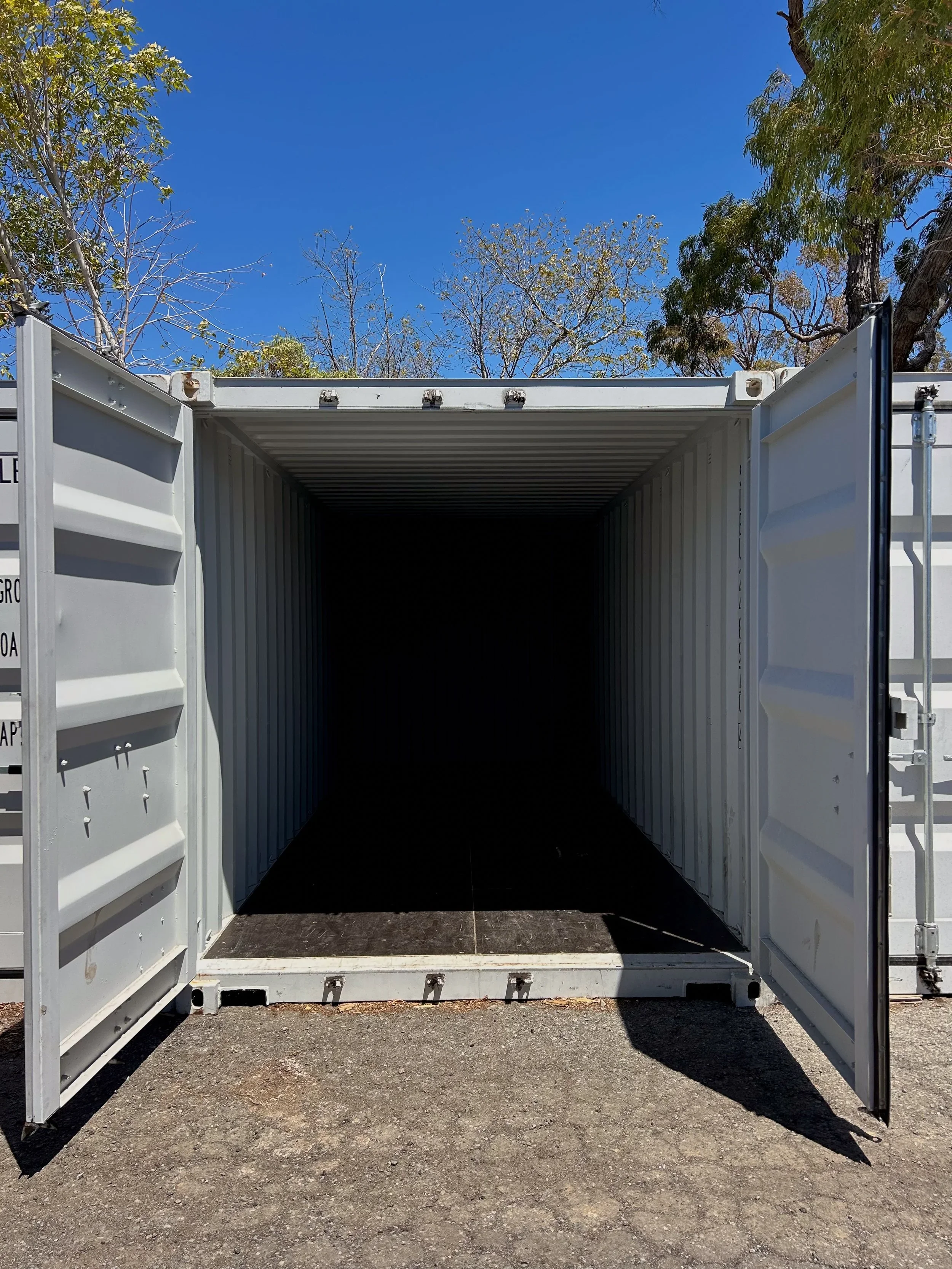 Open white shipping container on gravel ground with trees and blue sky in background.
