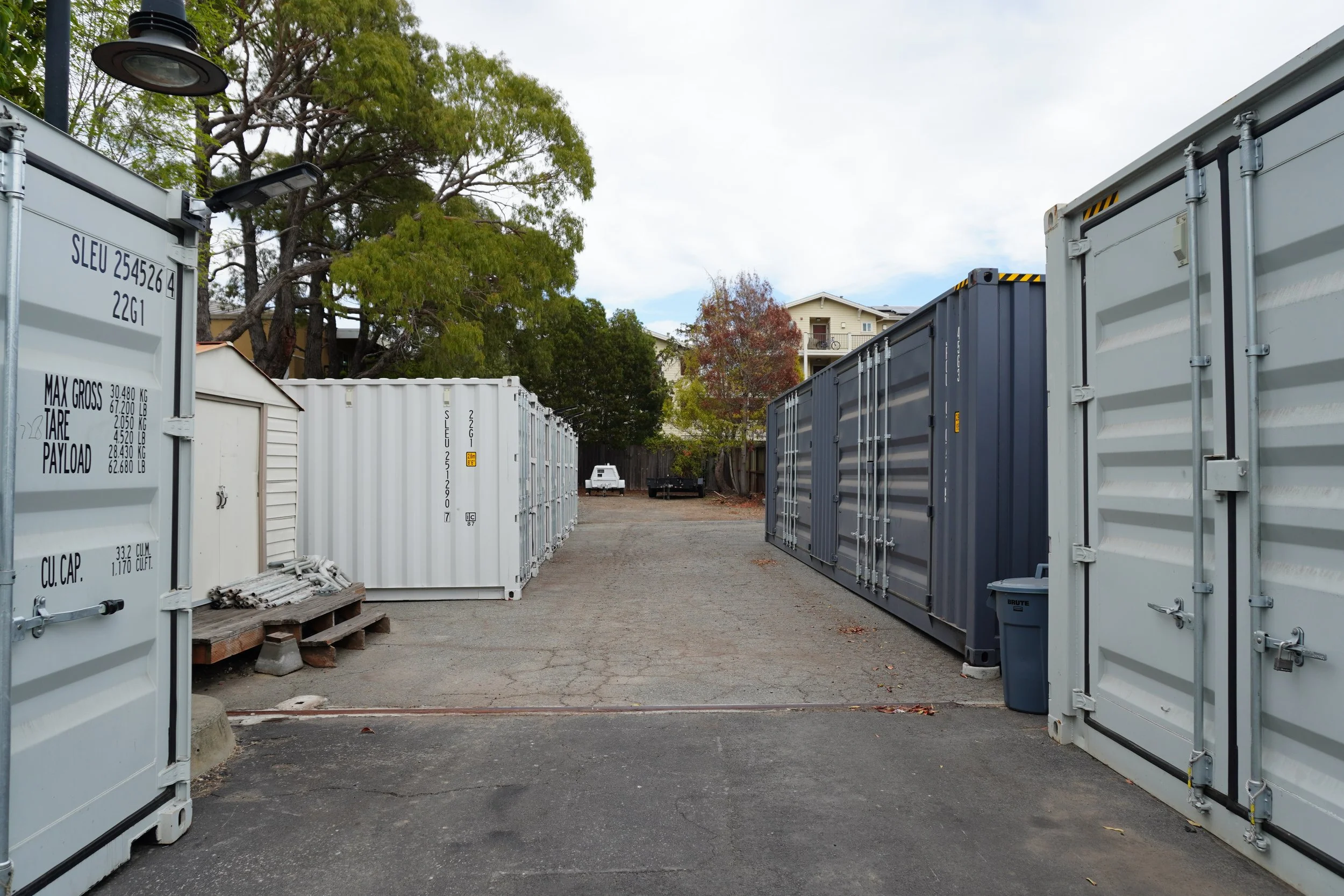 A storage yard with multiple shipping containers, some white and some gray, arranged in rows. There are trees and a residential building in the background, with cloudy sky overhead.