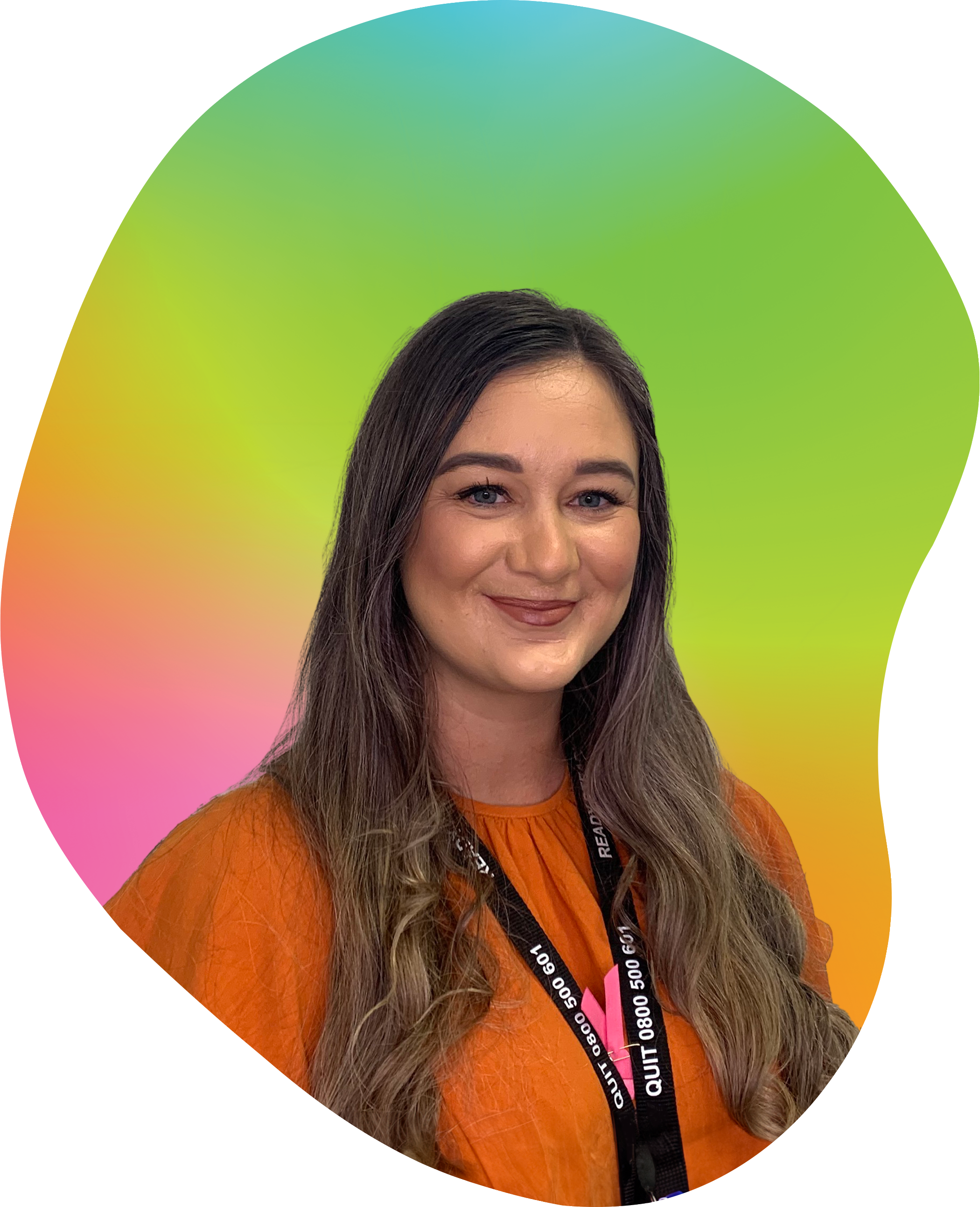 A woman with long brown hair and blue eyes smiling, wearing an orange top and a black lanyard with white text around her neck, standing in front of a colorful rainbow gradient background.
