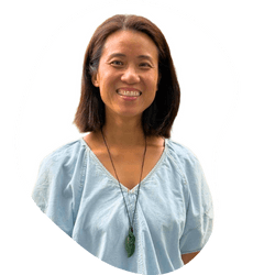 A woman smiling, wearing a light blue top and a necklace with a green pendant, standing against a white background.