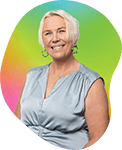 Smiling elderly woman with short white hair wearing a light blue blouse, standing against a colorful background with pink, green, and yellow hues.
