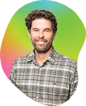 A smiling man with a beard and short hair, wearing a plaid shirt, standing in front of a colorful background.