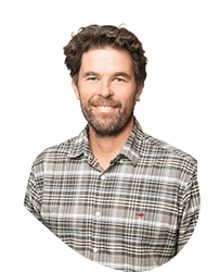 A smiling man with dark, curly hair and a beard, wearing a plaid button-up shirt.