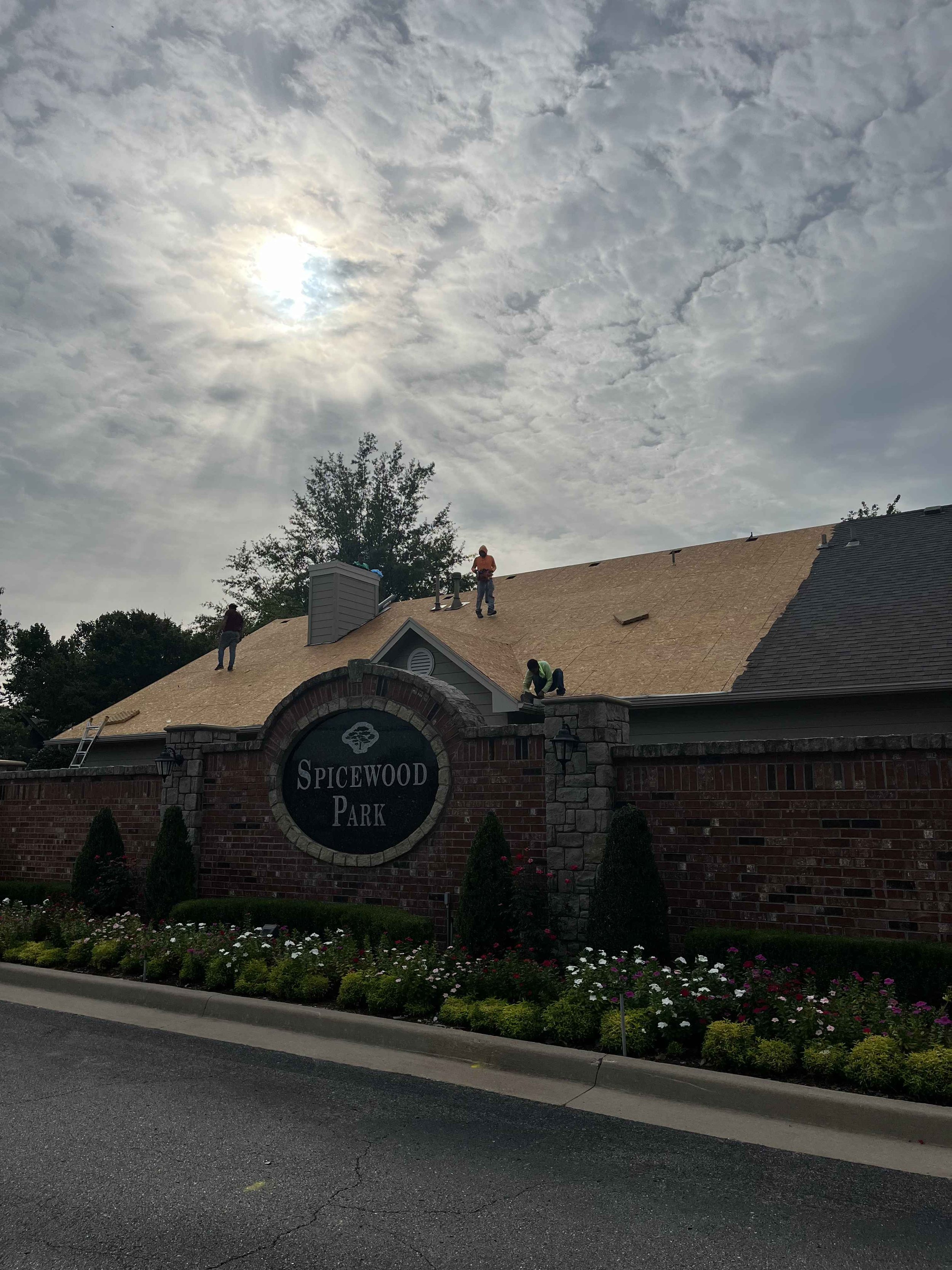 The entrance sign of Spicewood Park with a brick wall, flowers in front, and a roof with workers repairing it under a cloudy sky with the sun peeking through.
