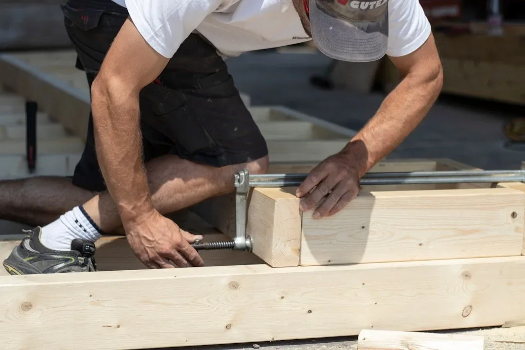 A person kneeling and using a clamp to hold a wooden block while working on a piece of wood in a workshop.