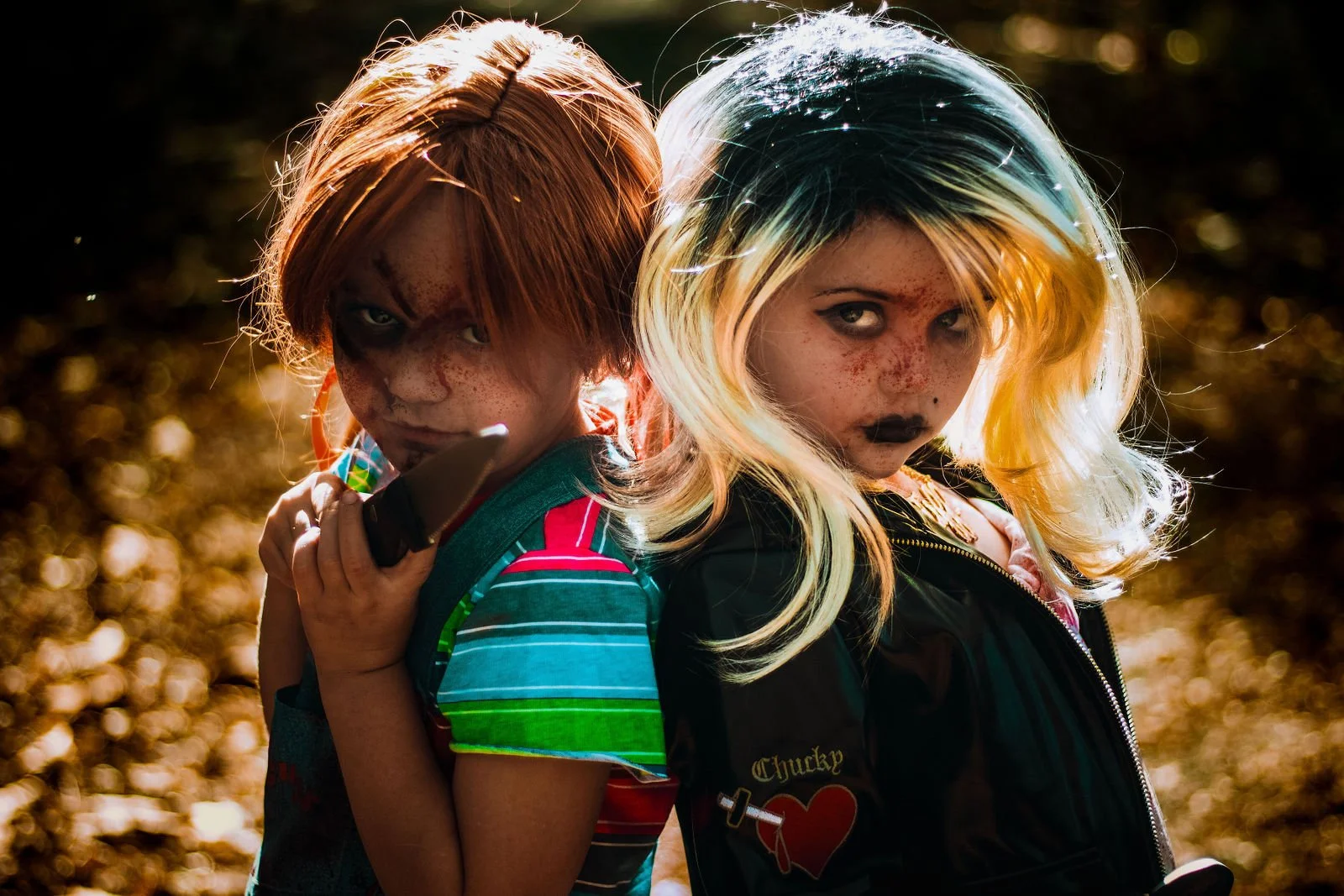 Two children with intense expressions pose back to back outdoors in sunlight, one with orange hair and the other with blonde hair with dark eyeliner and makeup.