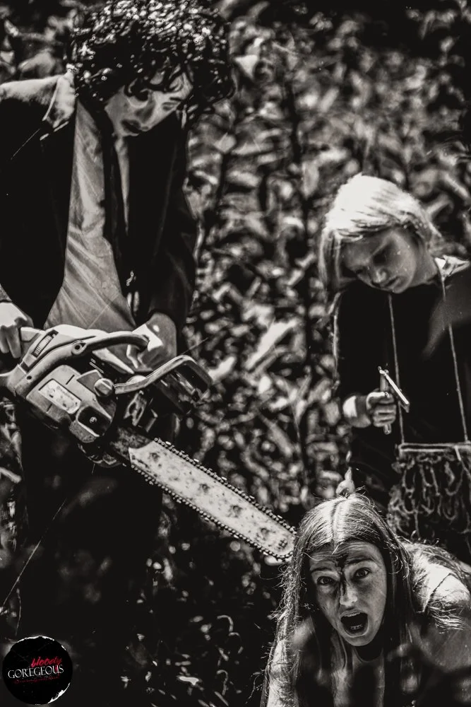 Black and white photo of three women outdoors, one lying on the ground with a surprised or fearful expression, holding a chainsaw, with two other women standing nearby, one holding a tool, all surrounded by dense foliage.