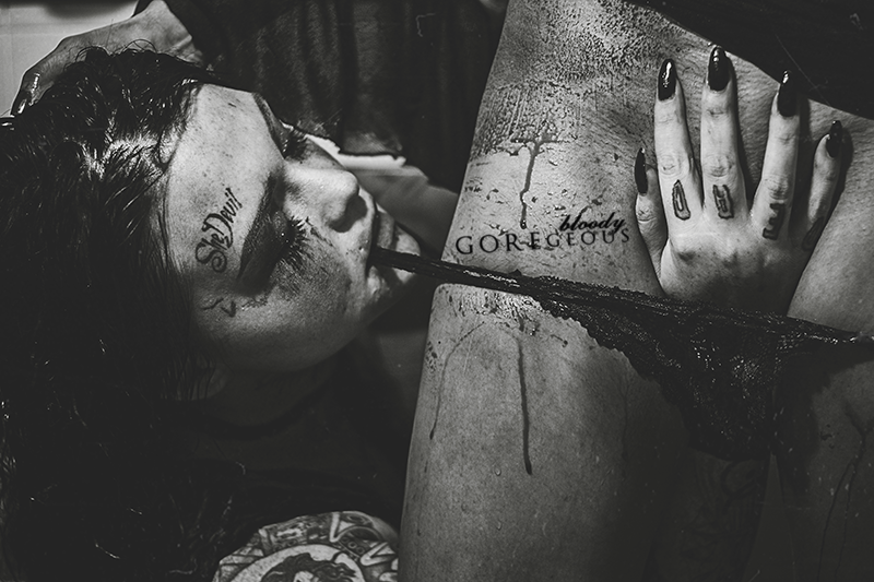 Black and white photo of a woman with tattoos, lying down and smoking a cigarette. The woman's face has tattoos, including the words 'Slut' and 'Gorgeous.'