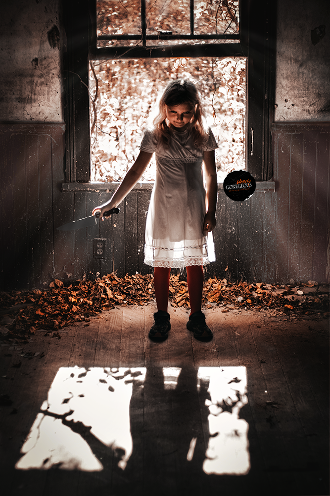 A young girl in a white dress standing in front of a window with sunlight creating shadows on the floor, holding a knife in one hand and a black balloon in the other, in a dark, abandoned room with dead leaves on the ground.