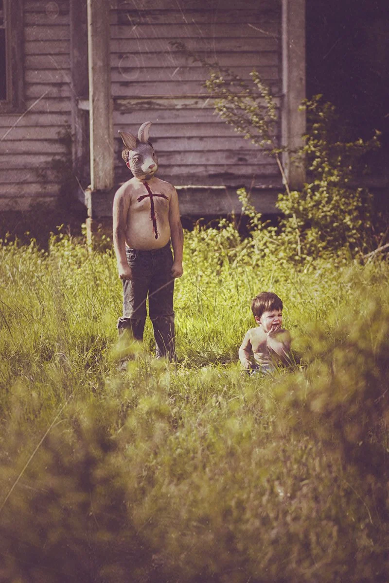 A child in a rabbit mask and body paint resembling a crucifix stands next to a crying toddler in an overgrown outdoor area near an old wooden building.