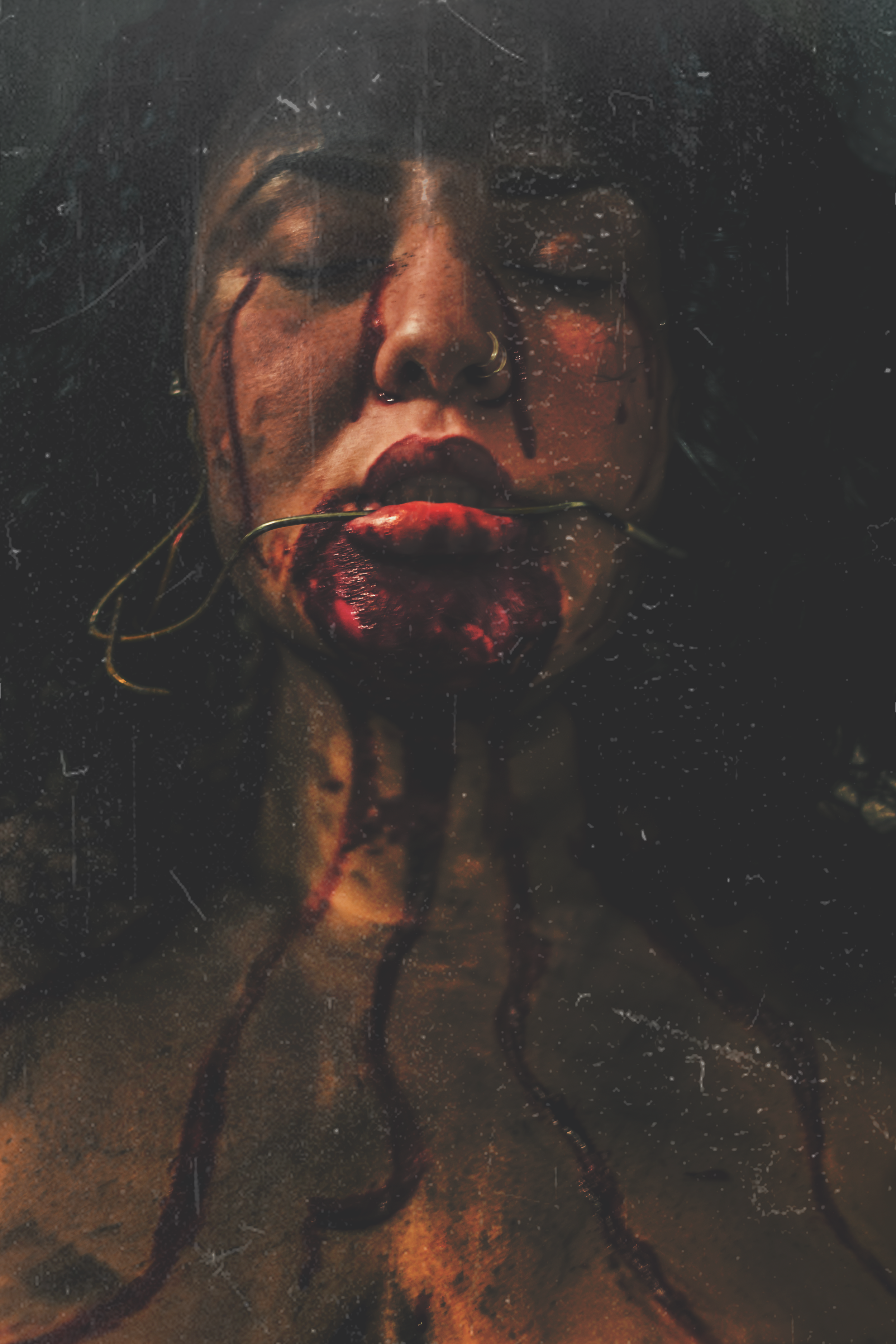 Woman with long dark hair and closed eyes, with blood and a plant in her mouth, and blood on her face, in a dark and gritty setting.