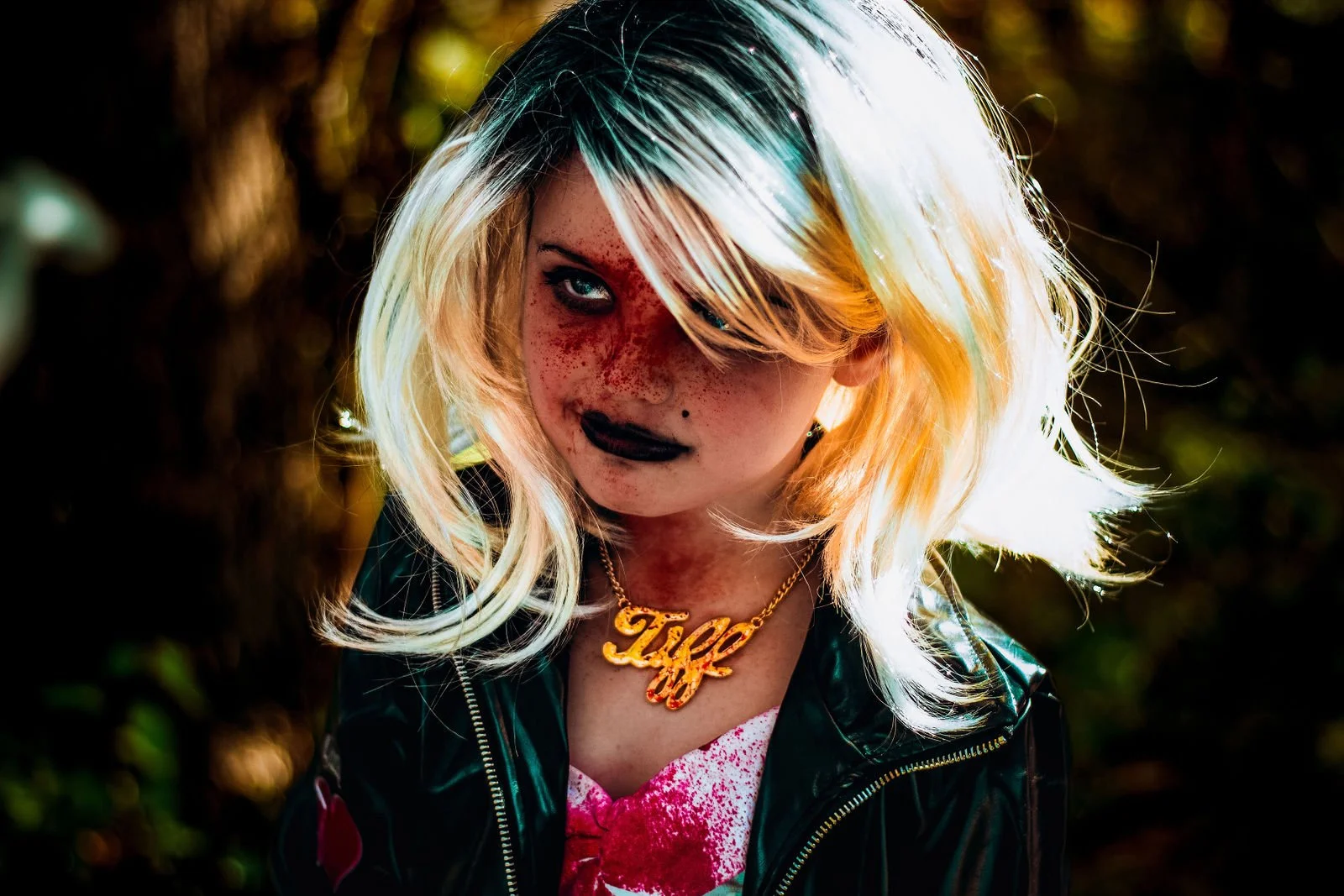 A woman with platinum blonde hair with dark roots, wearing dark makeup and a gold necklace that says "Tey". She has a serious expression, freckles, and is dressed in a black leather jacket over a pink top with red splashes.