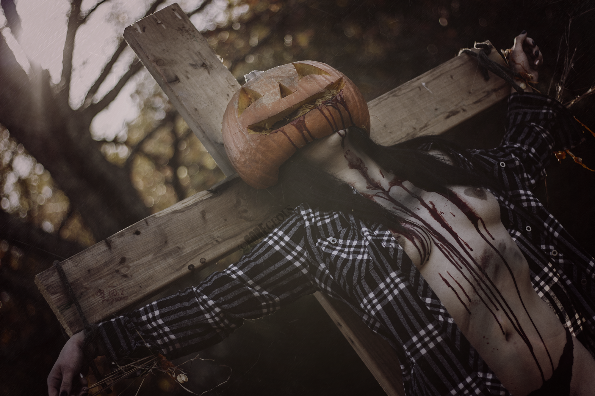 A Halloween-themed decoration featuring a carved pumpkin with a face, mounted on a wooden cross or structure, with a figure underneath resembling a zombie or Frankenstein character with blood on its chest, dressed in a plaid shirt, outdoors with blur