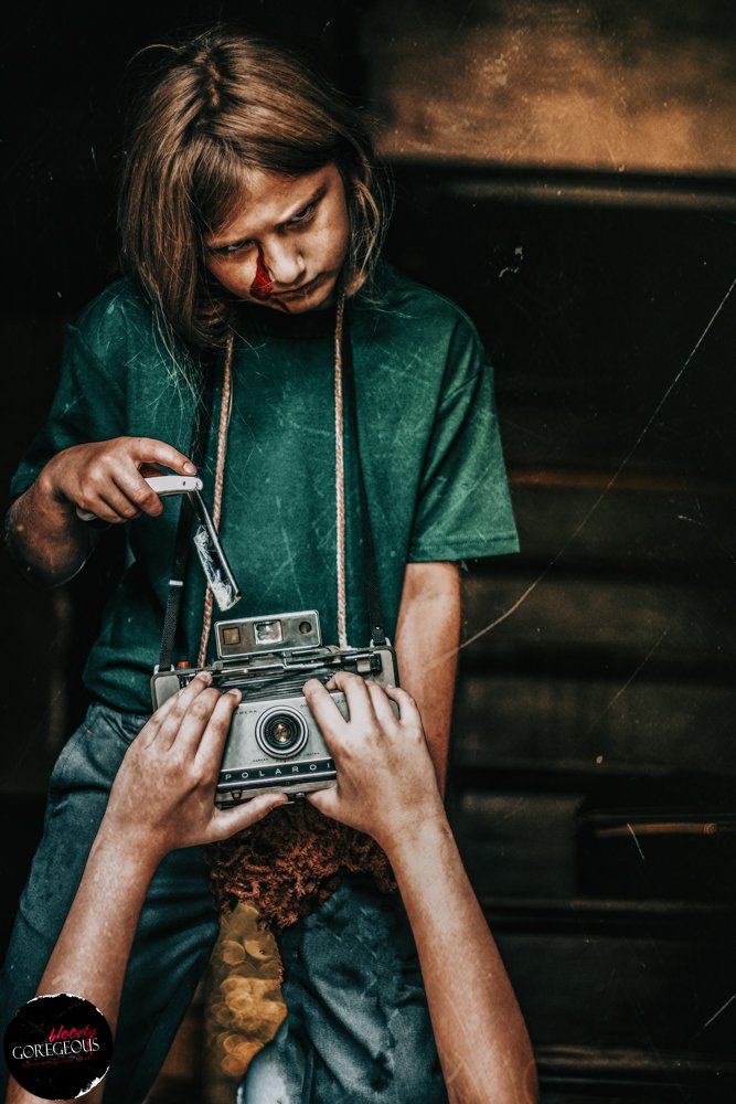 A young girl with a nosebleed looks down at an old camera held by another person's hands, in a dark setting.