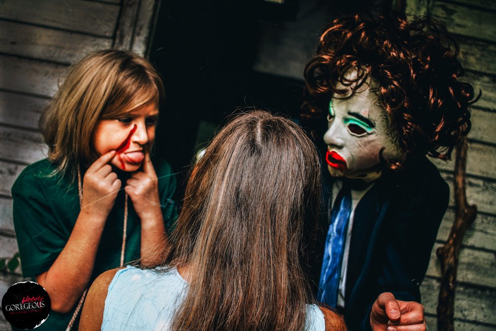 A person in a horror mask with red curly hair and makeup is speaking to two women, one with long blonde hair and the other with brown hair, in a dimly lit setting with wooden walls.